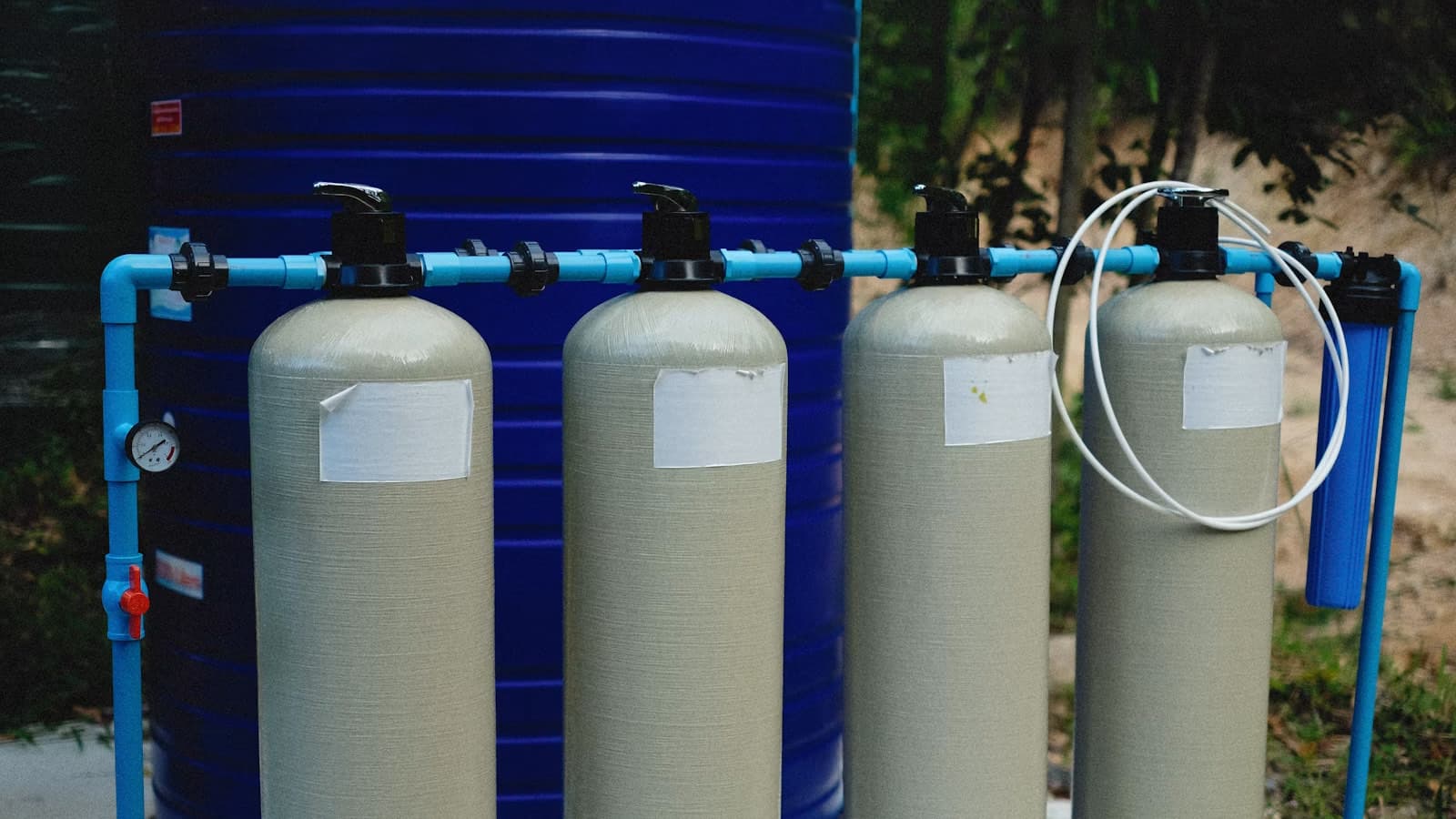 Four cylindrical water-softener resin tanks with blank labels, connected by blue piping in front of a large blue storage tank.