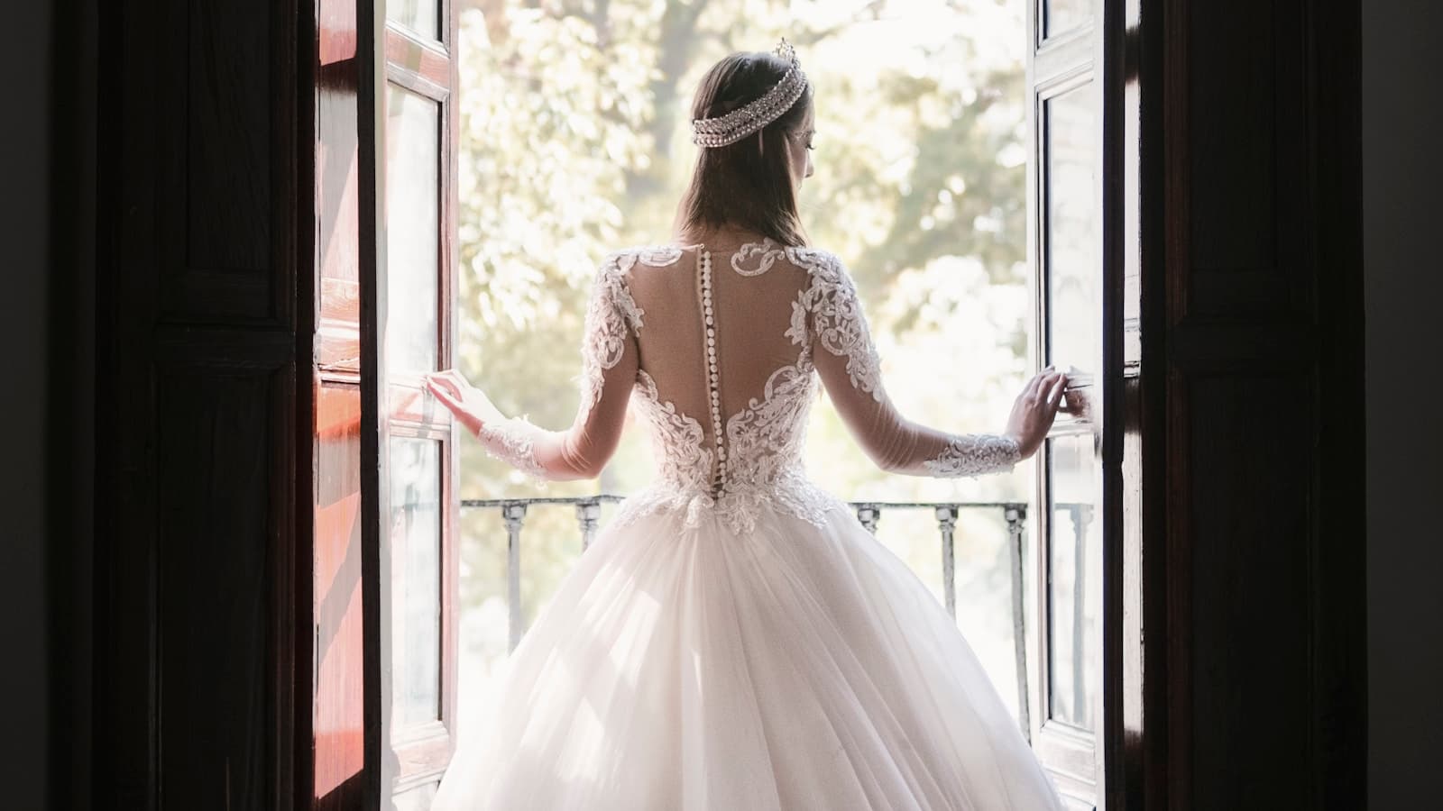 A bride in a lace wedding gown and tiara seen from behind, standing at open French doors overlooking a garden.
