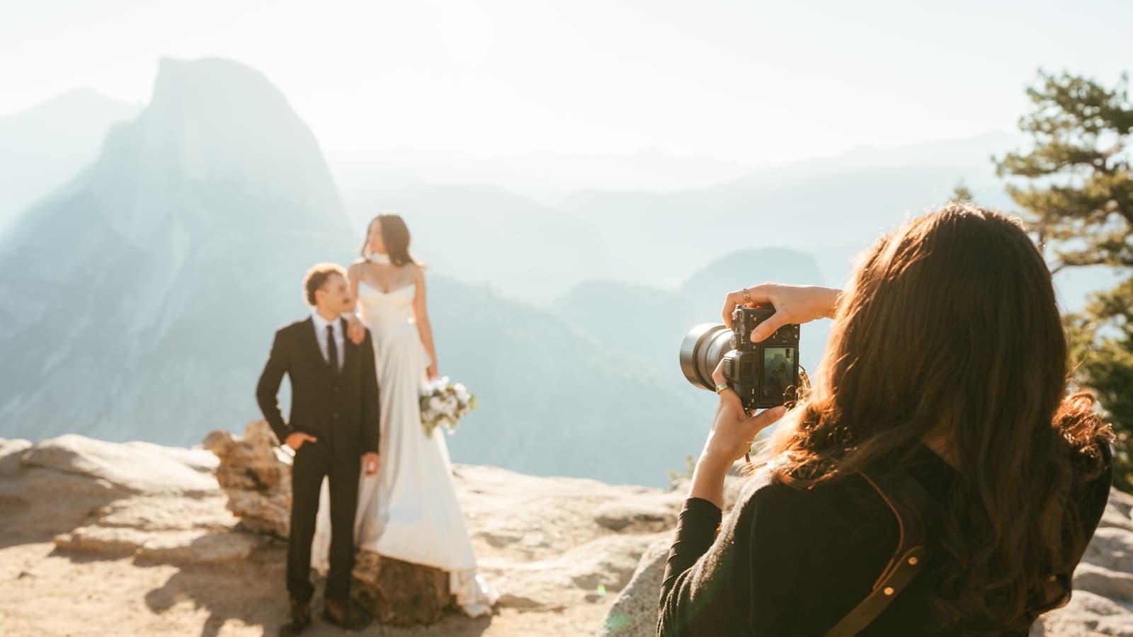 Bride and groom posing for a photographer on a mountain overlook.