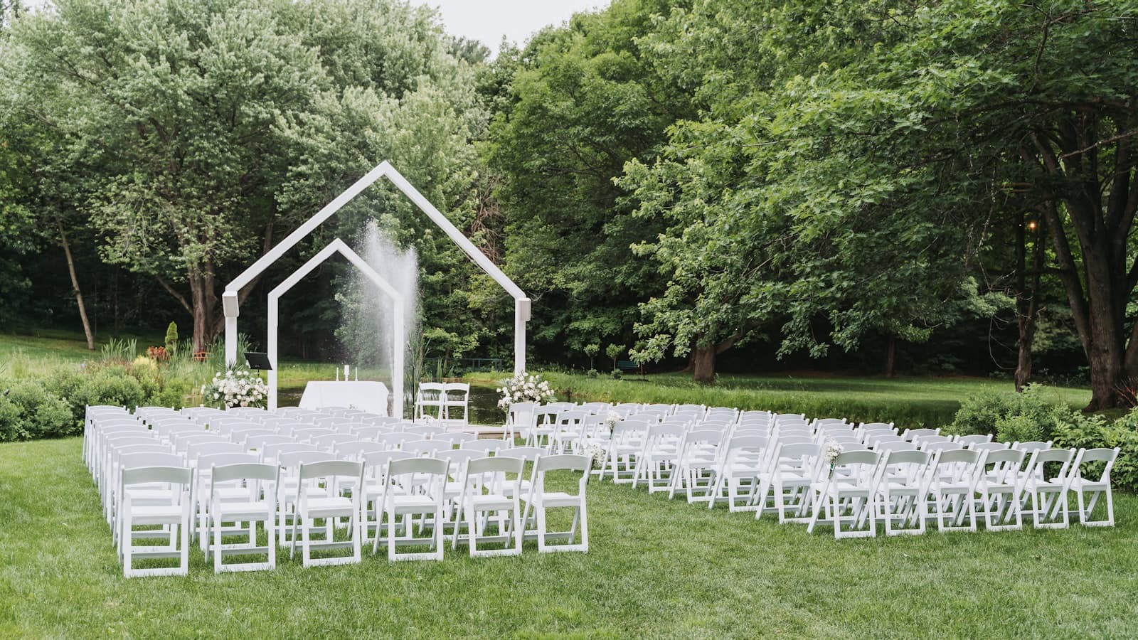 Rows of white folding chairs facing a house-shaped wedding arch and fountain on a grassy lawn surrounded by trees, no people visible.