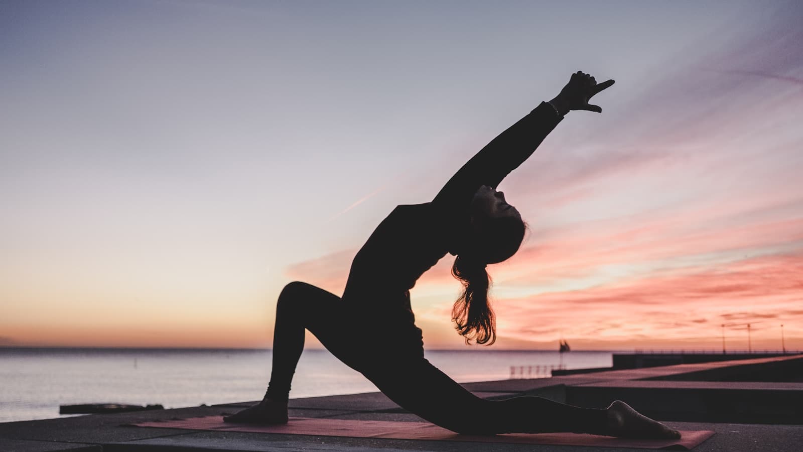 Silhouette of a woman doing a yoga pose outdoors at sunset by the water.