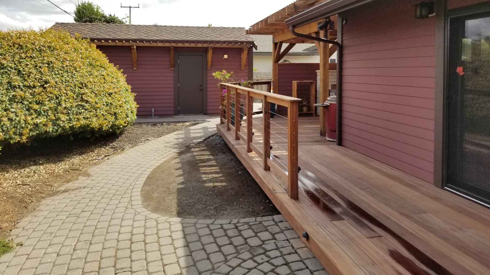 A wooden wheelchair ramp with a cable railing curves from a paver walkway up to a covered deck at the back of a house.