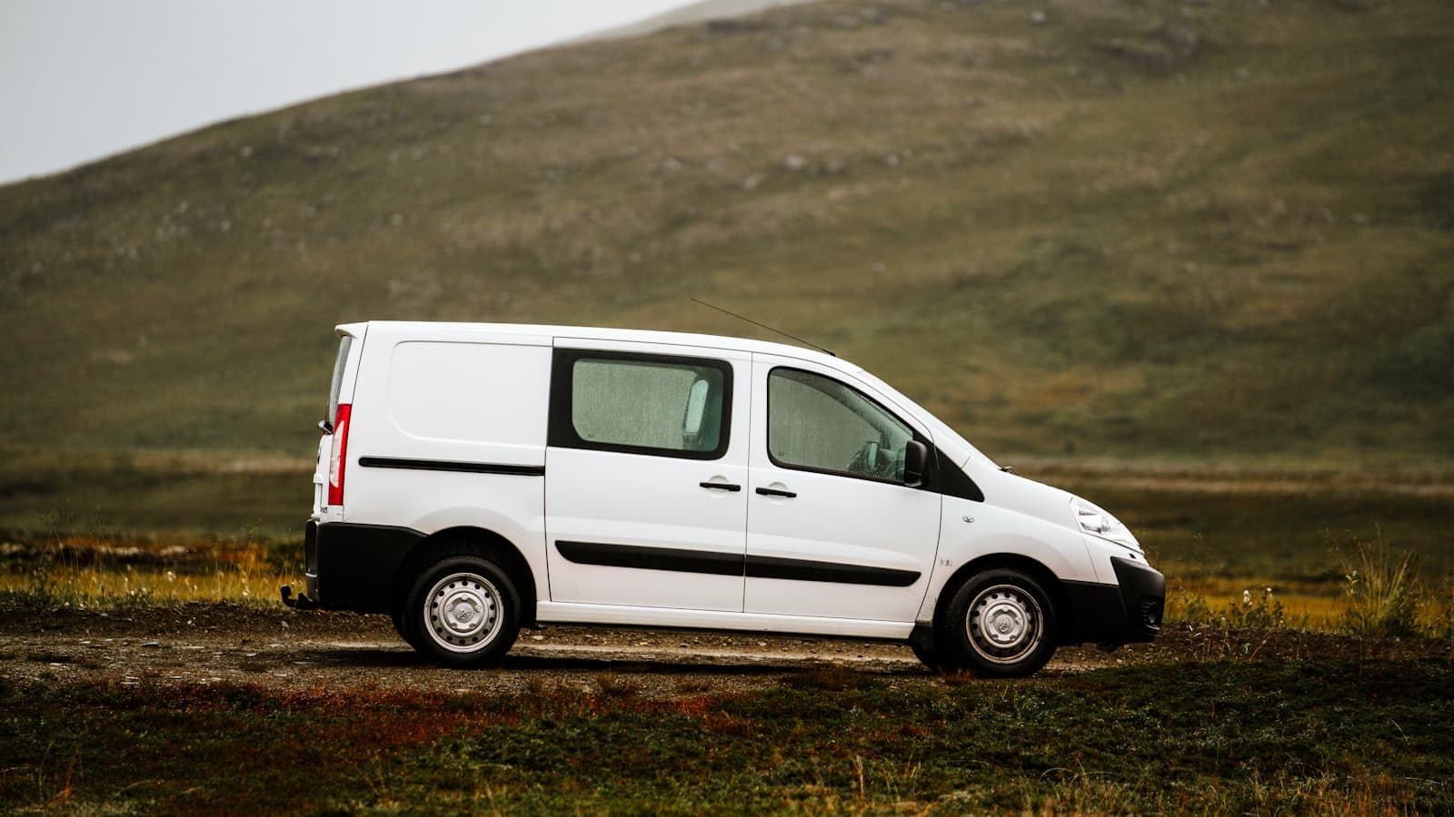 A white cargo van in side profile parked on a dirt track in open countryside, with no visible plate, logo, or people.