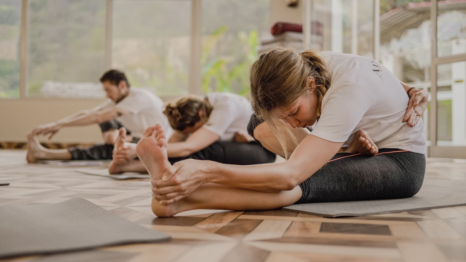 A yoga instructor assisting a student into a seated forward fold, other students practicing nearby.