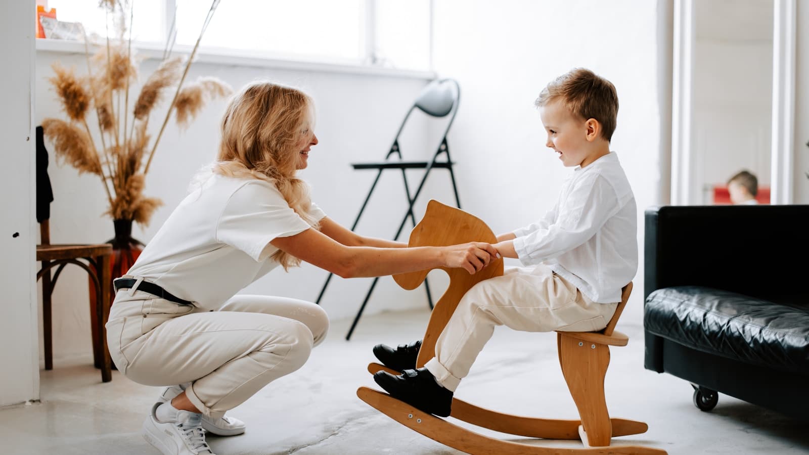 A woman kneeling to play one-on-one with a young boy on a wooden rocking horse.