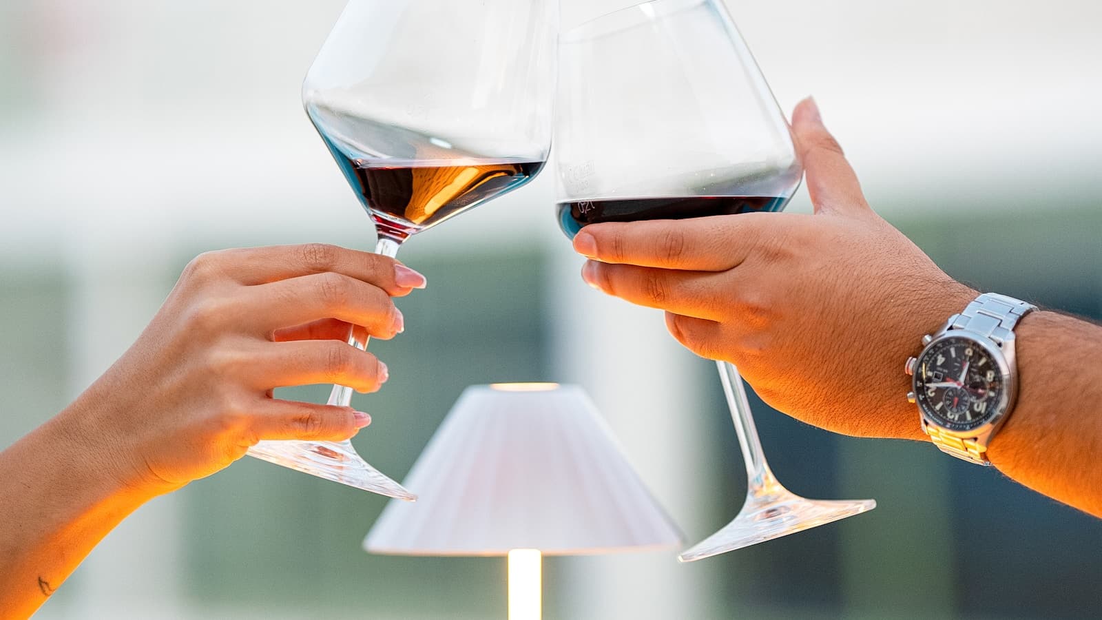 Close-up of two hands clinking wine glasses at a warmly-lit rooftop setting.