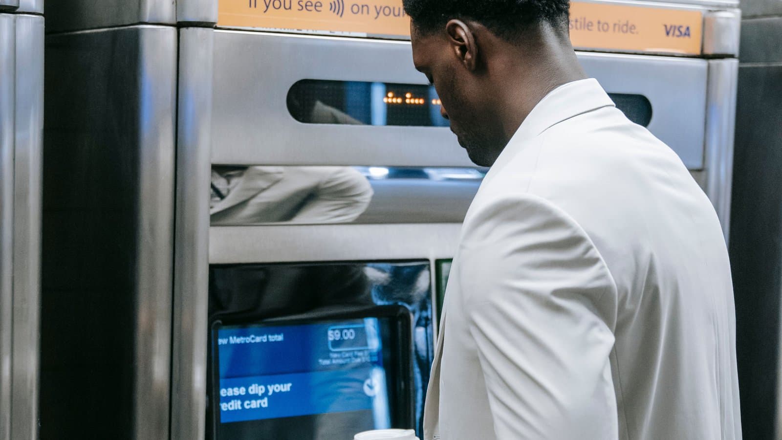 A man in a light suit holding a coffee cup while using a bank ATM machine.