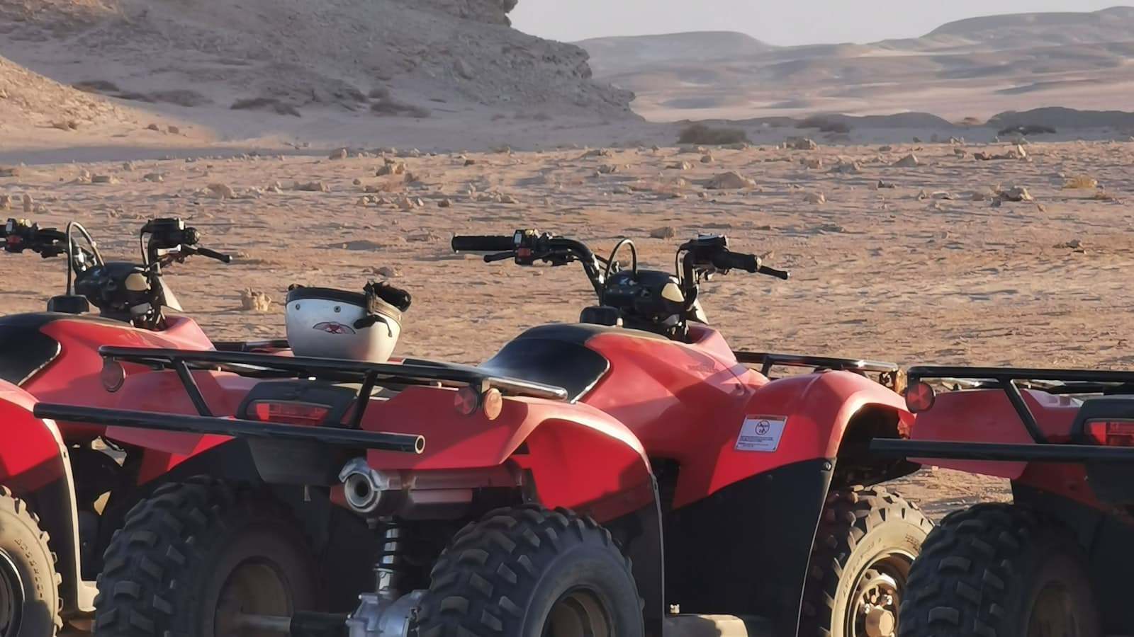 Rear view of two red ATVs parked side by side in a desert with helmets resting on the cargo racks, no faces, plates, or readable brand text visible.