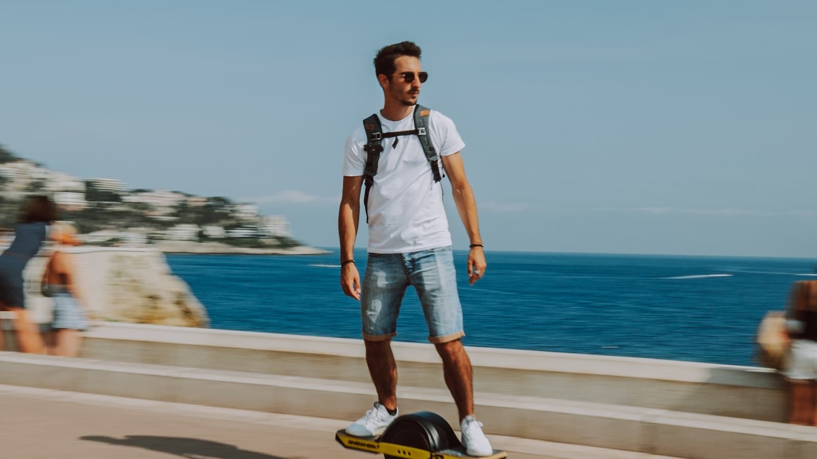 A man rides an electric skateboard along an oceanfront promenade.
