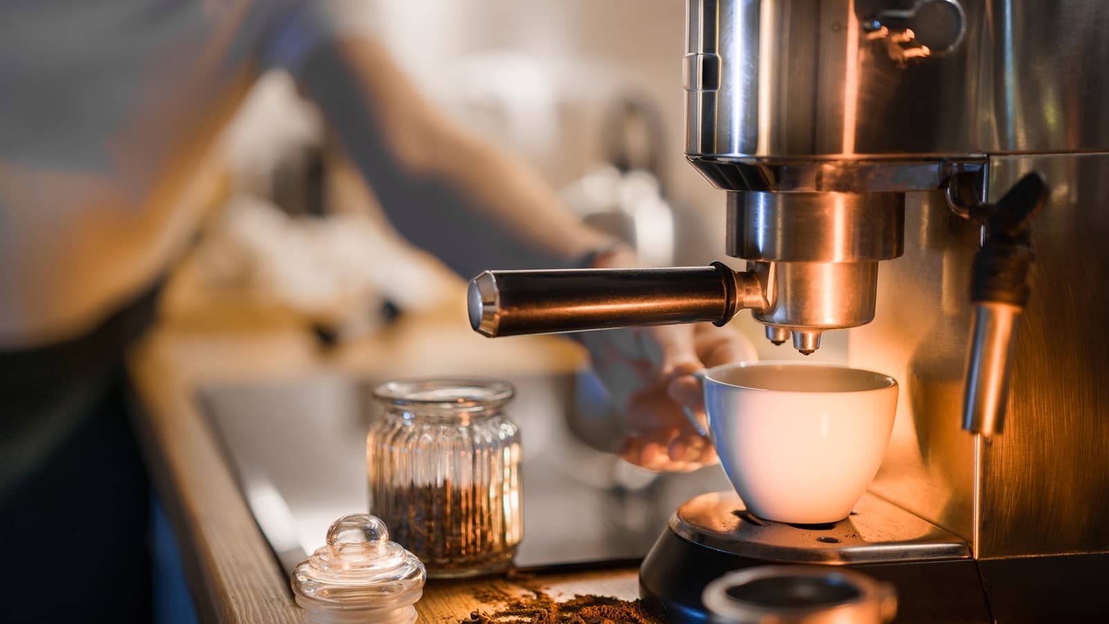 A stainless steel espresso machine pulling a shot into a white cup, with a blurred person in an apron in the background.