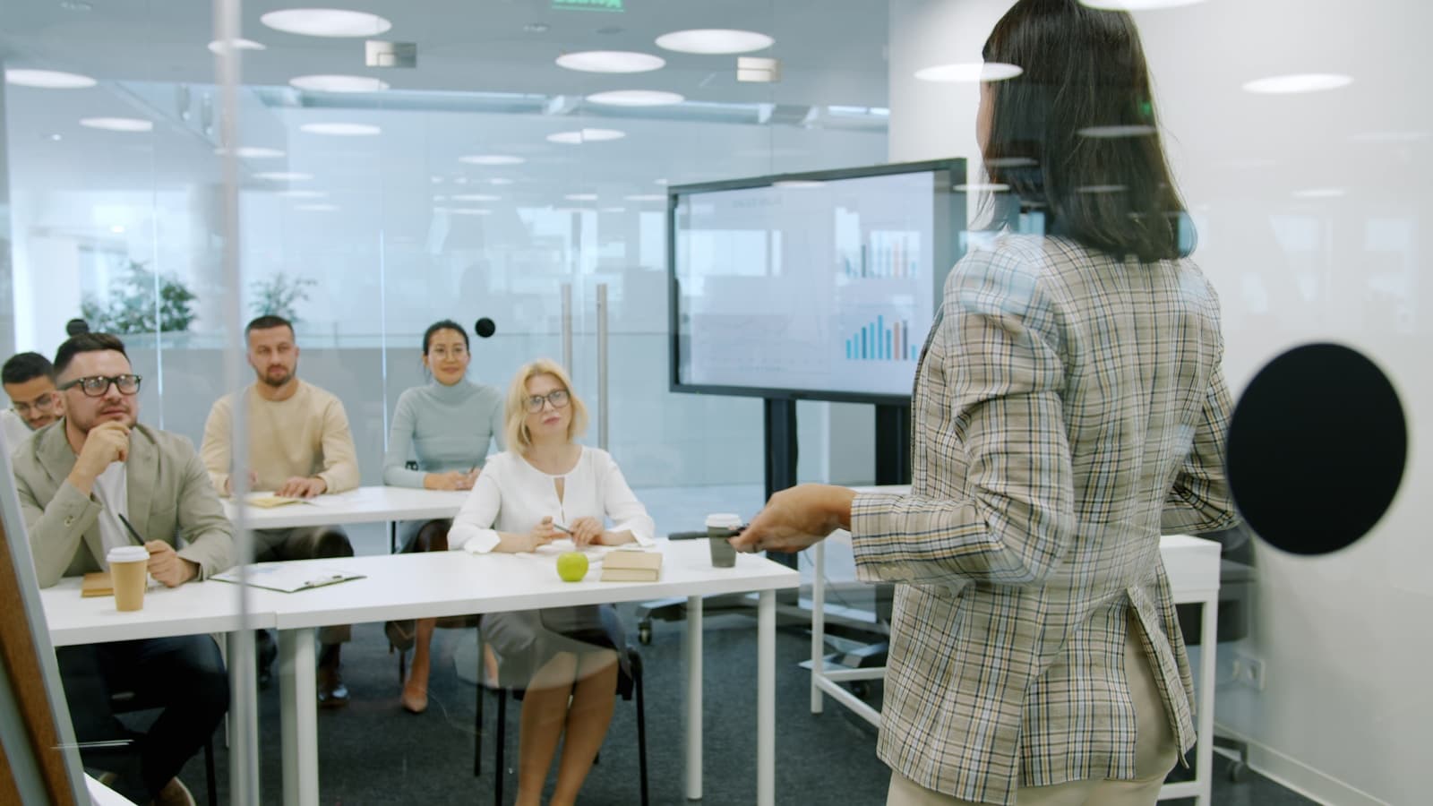 A presenter walking a small group through bar-chart data in a glass-walled meeting room.
