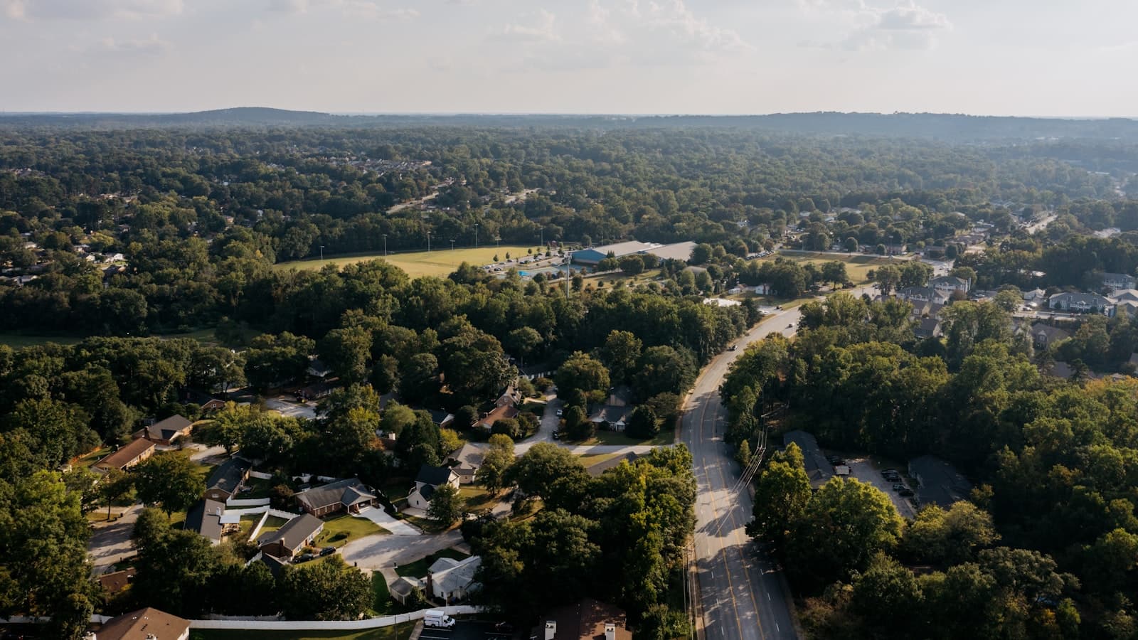 An aerial view of a tree-lined suburban residential neighborhood with winding streets and houses, with no people or readable text visible.