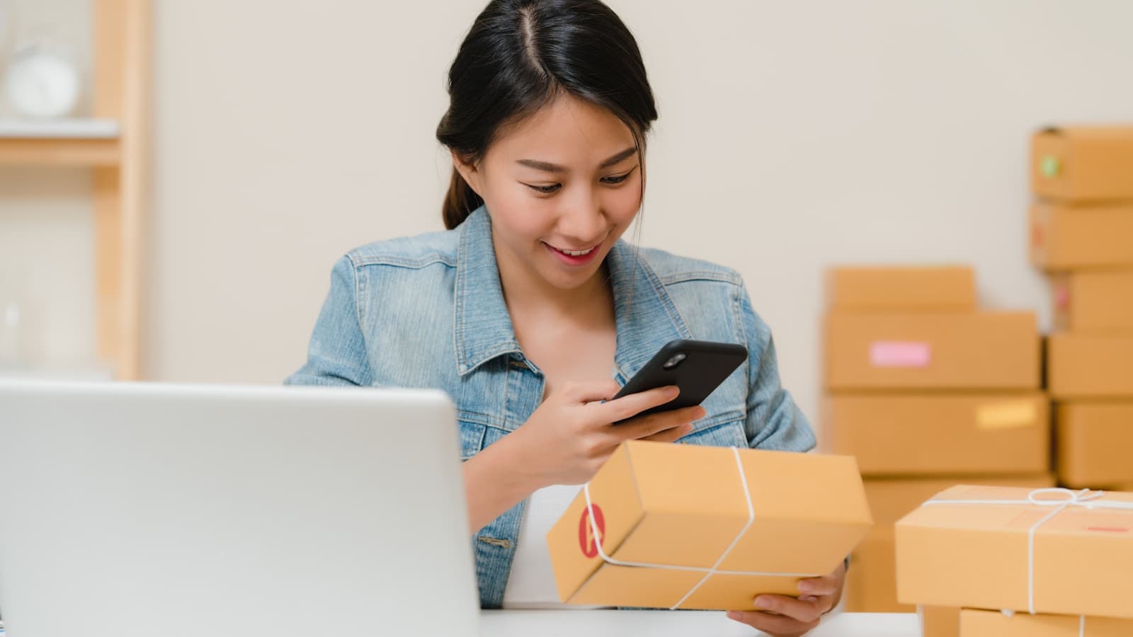 A smiling woman checking a shipping label on her phone while packing boxes.