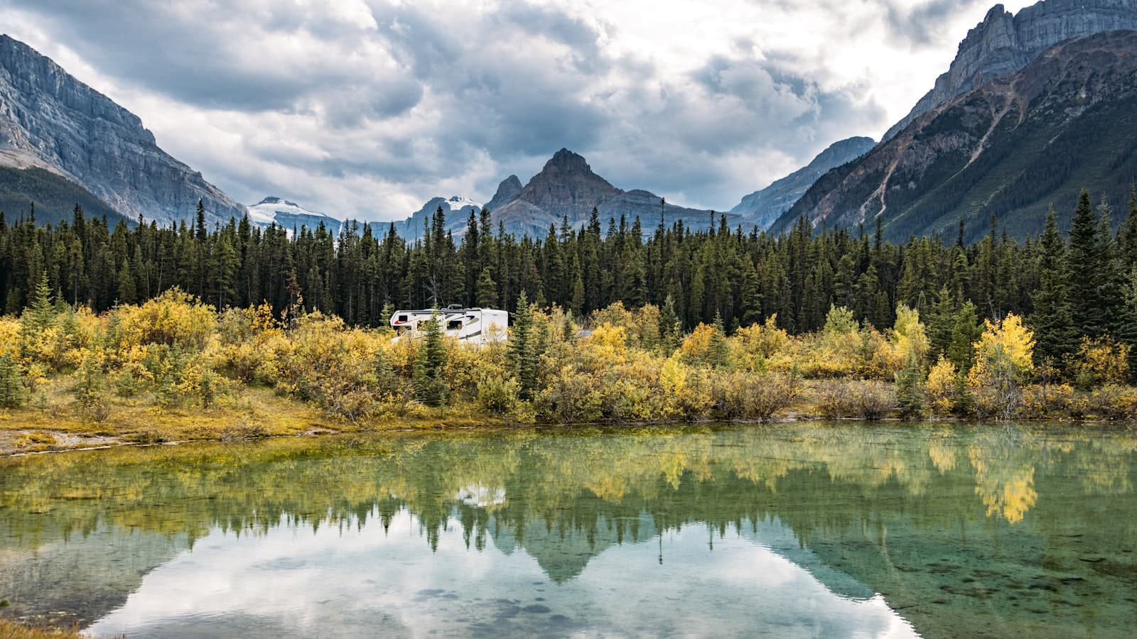 A white Class C motorhome parked among yellow autumn shrubs and evergreen trees, framed by mountains and reflected in a calm lake.