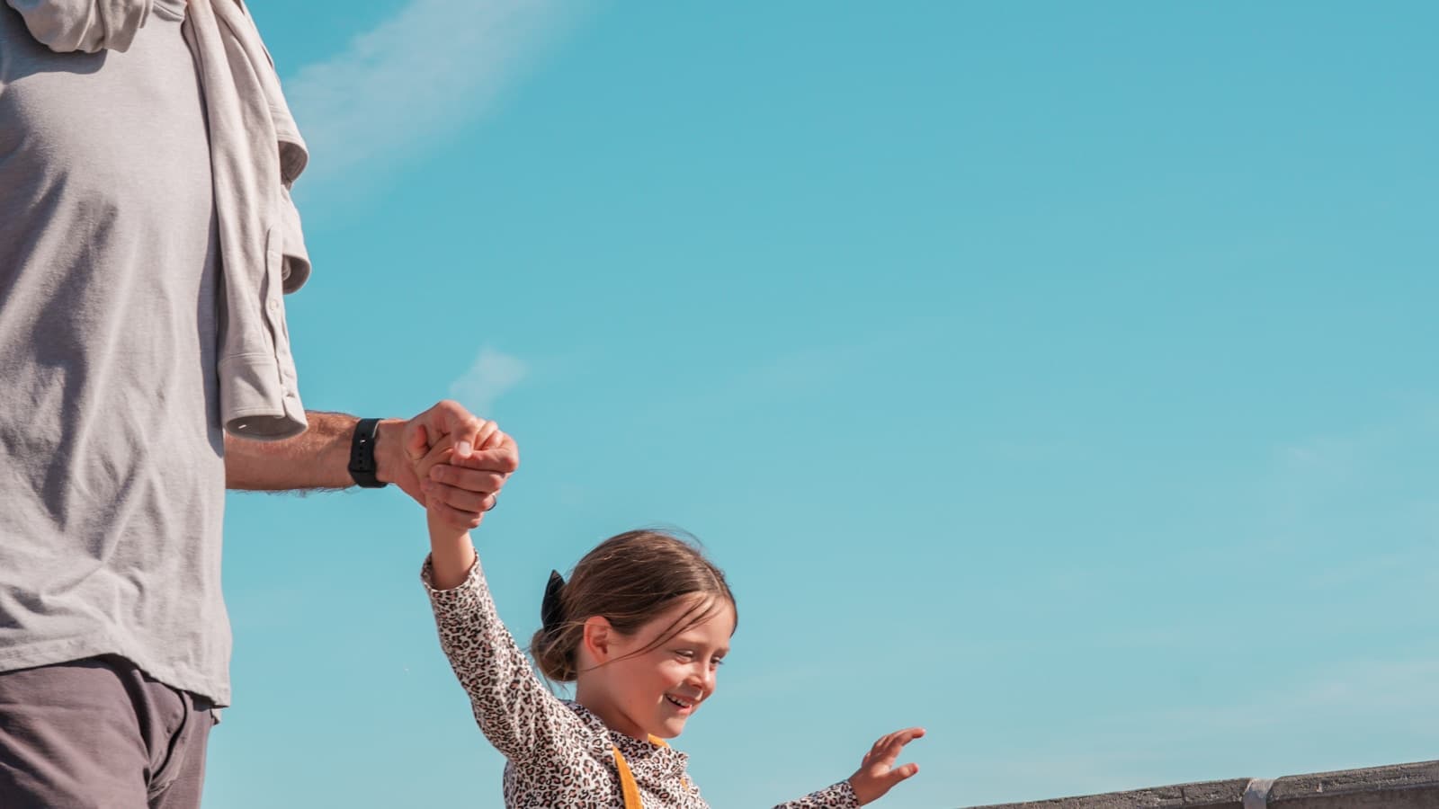 A father and daughter holding hands on a walk, bright blue sky.