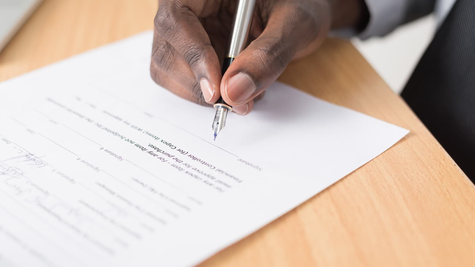 Close-up of a hand signing a printed contract with a fountain pen on a wooden desk, no face or legible text visible.