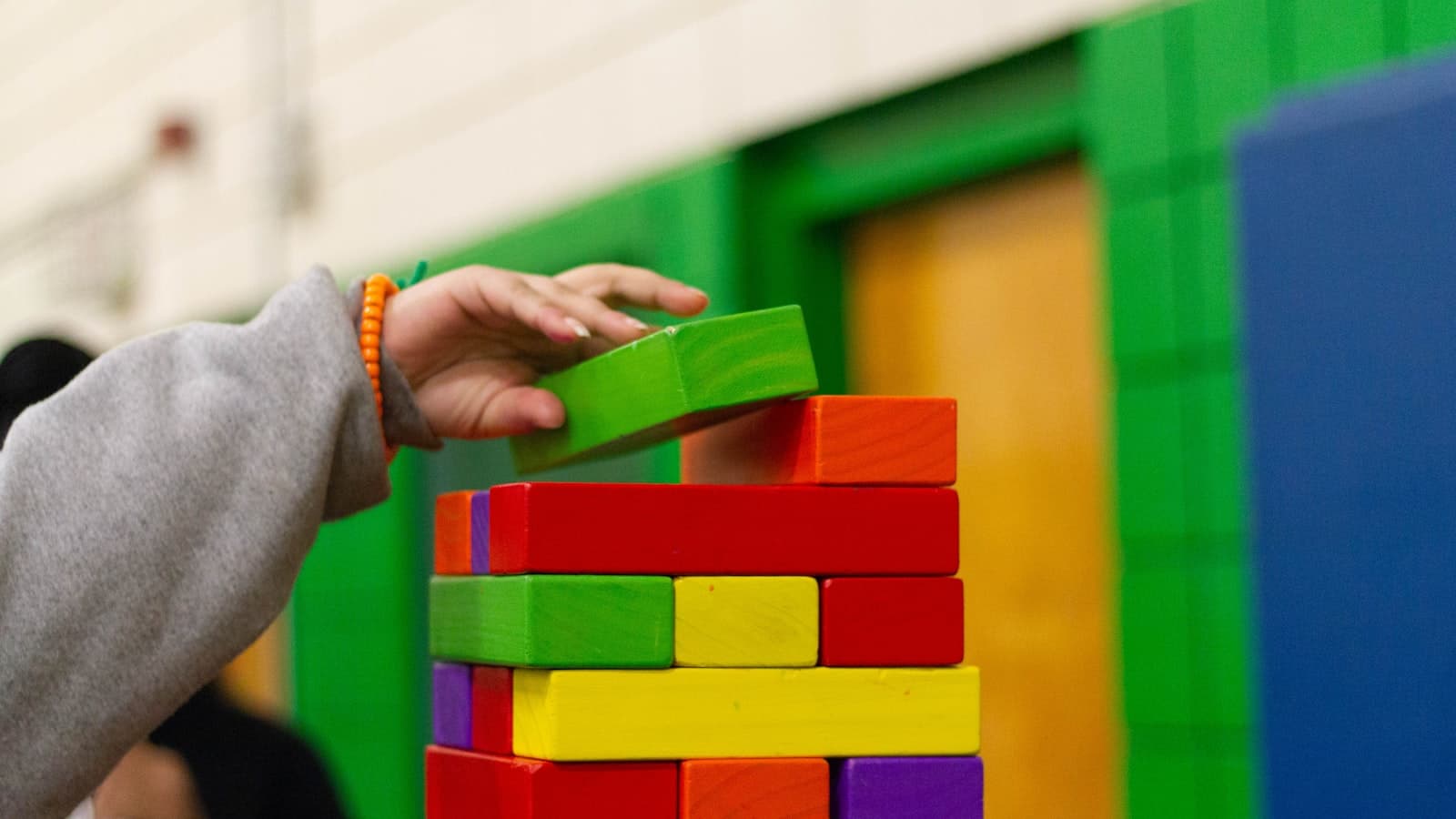 A child's hand stacking colorful wooden blocks with a blurred, unidentifiable figure in the background; no readable text or logos.