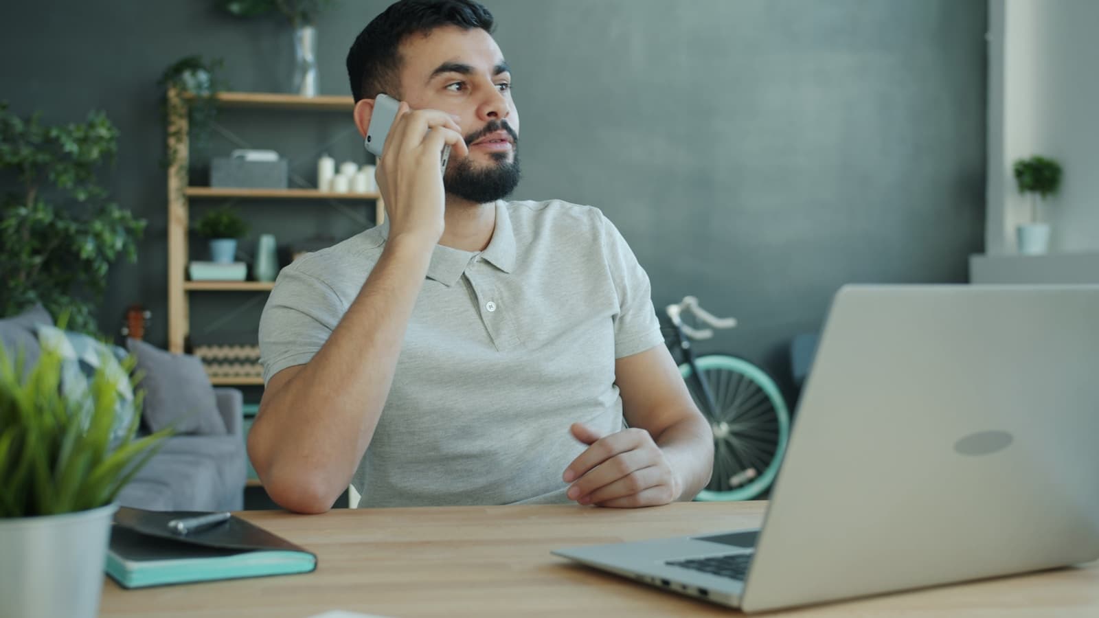 A man at a desk on a phone call next to an open laptop, calm and serious tone.