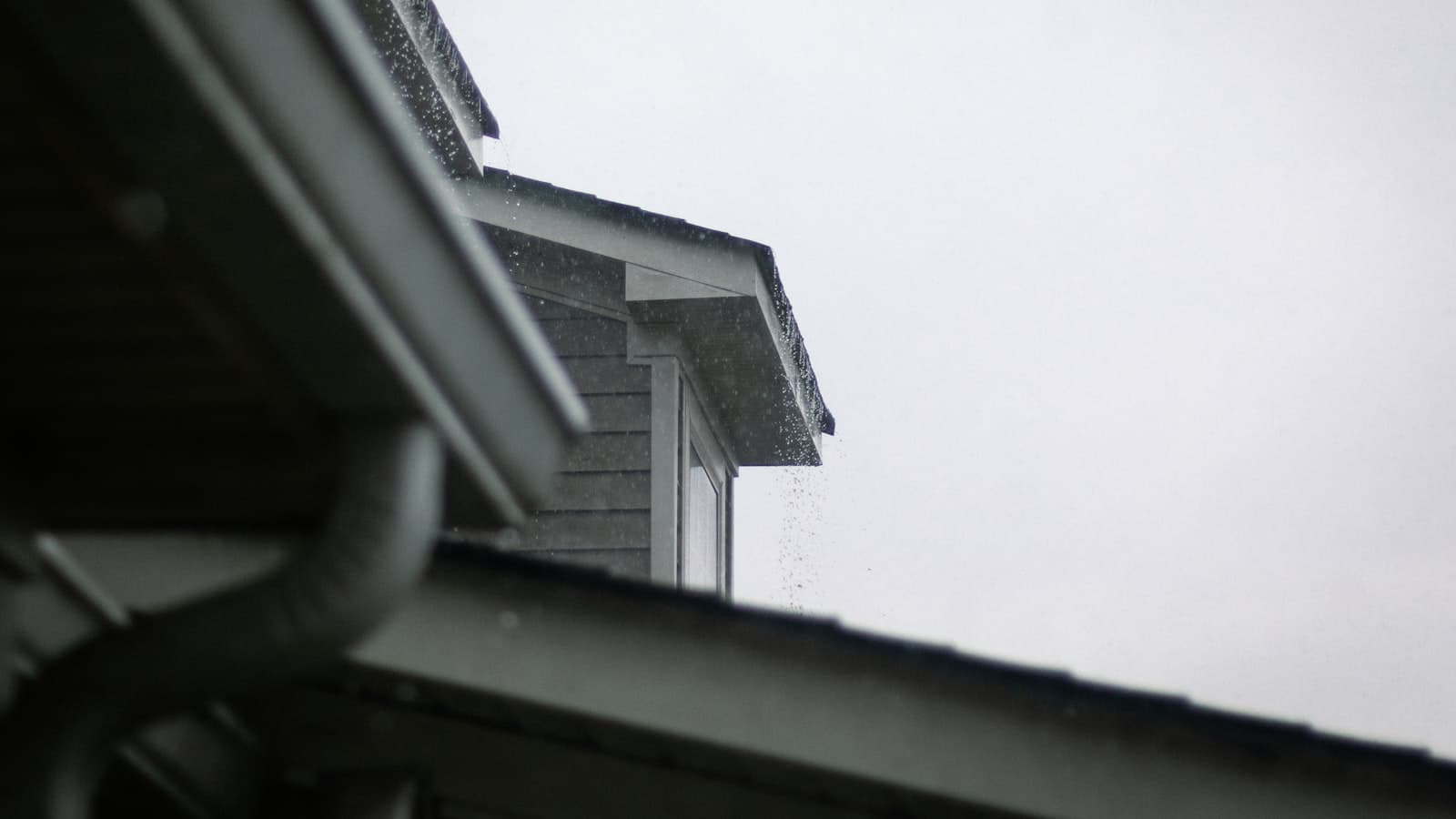 A close-up of a house roofline with a rain gutter and downspout under an overcast sky with light rain visible.