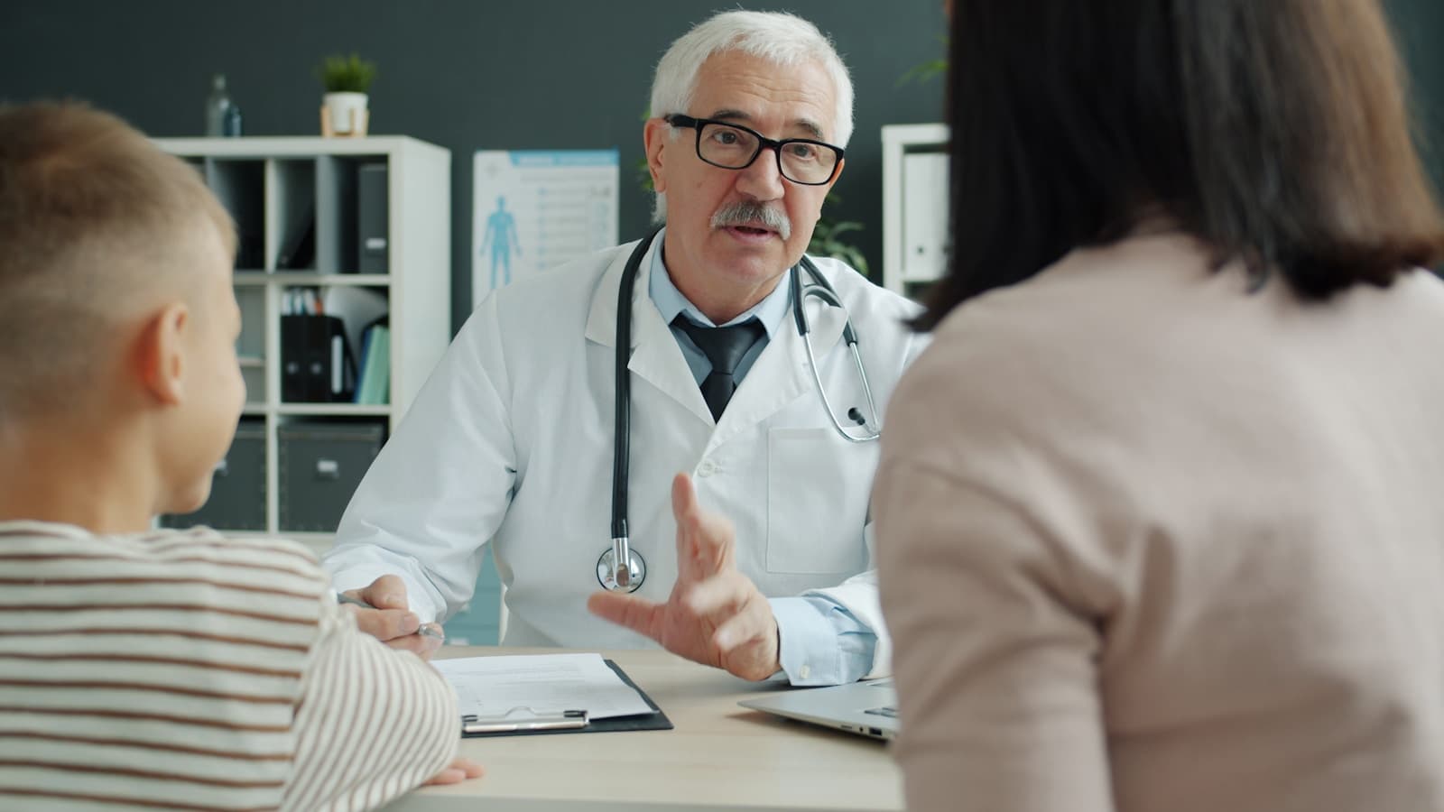 A senior doctor in a white coat consulting with a mother and her young son in a clinic office.