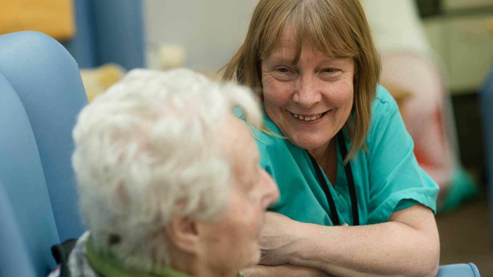 A nurse in scrubs leaning close and smiling with an elderly woman seated in a chair.
