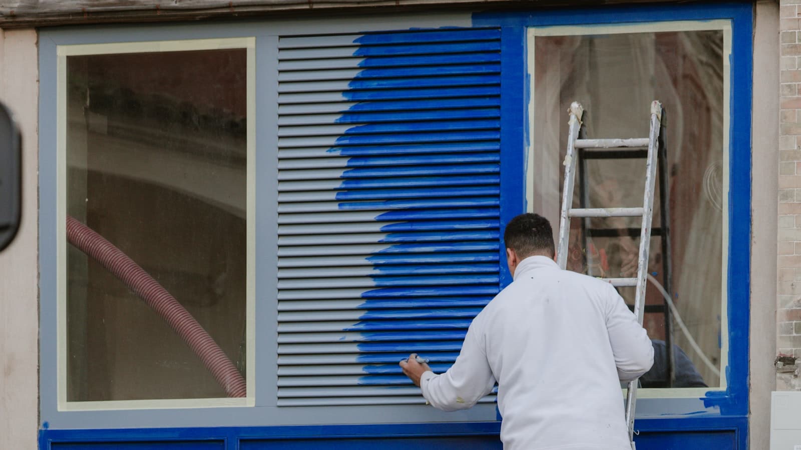 A person on a ladder painting a window shutter and frame blue on a house exterior with a taped-off window and stucco wall.