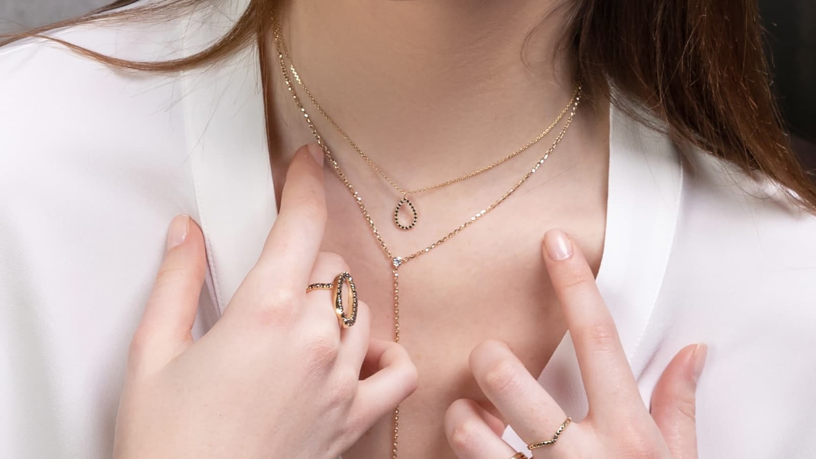Close-up of hands adorned with rings and a bracelet near a necklaced neck/chin; no identifiable face.