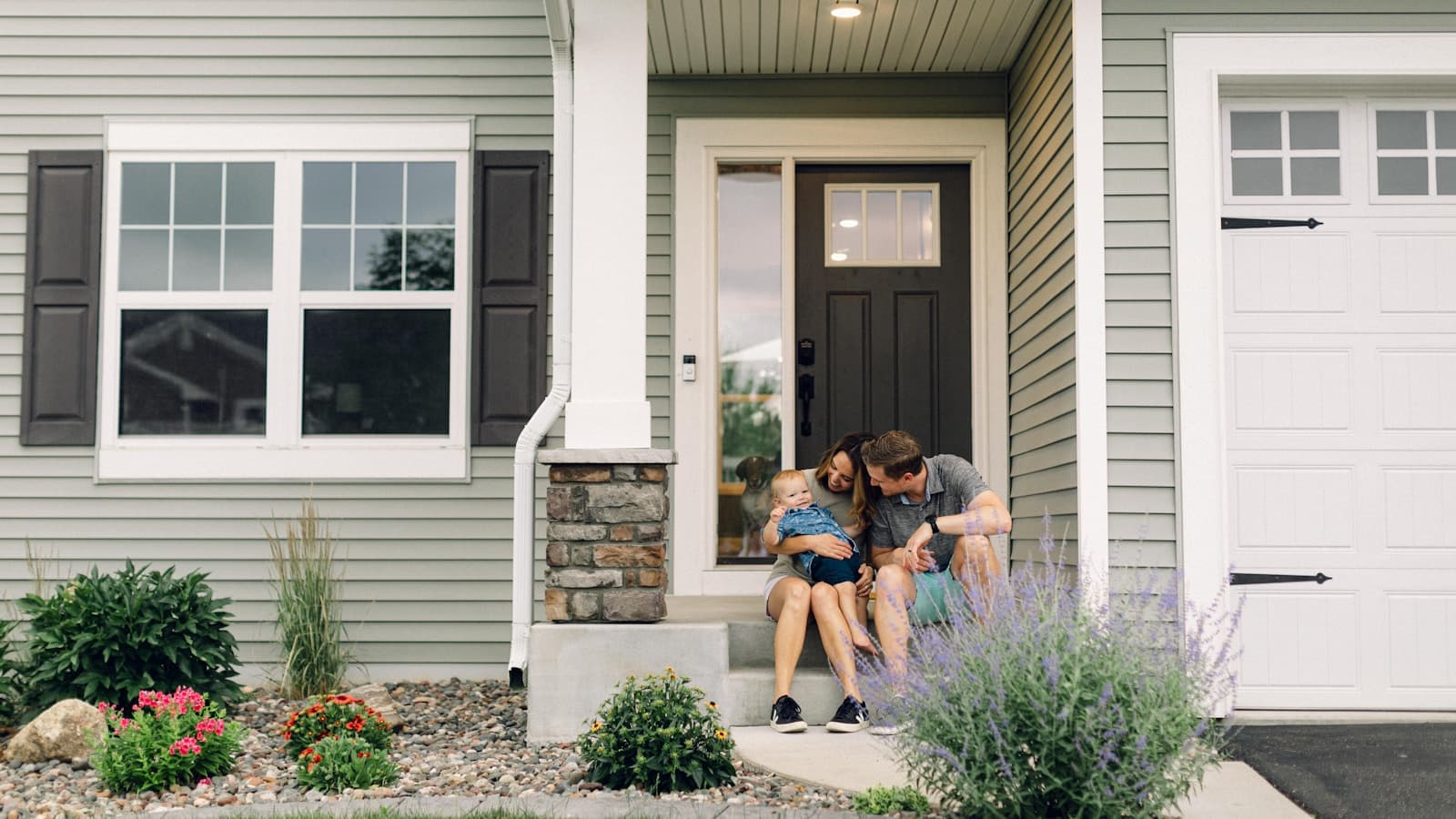 A young family with a toddler sitting together on the front steps of their house, a dog visible through the door behind them.