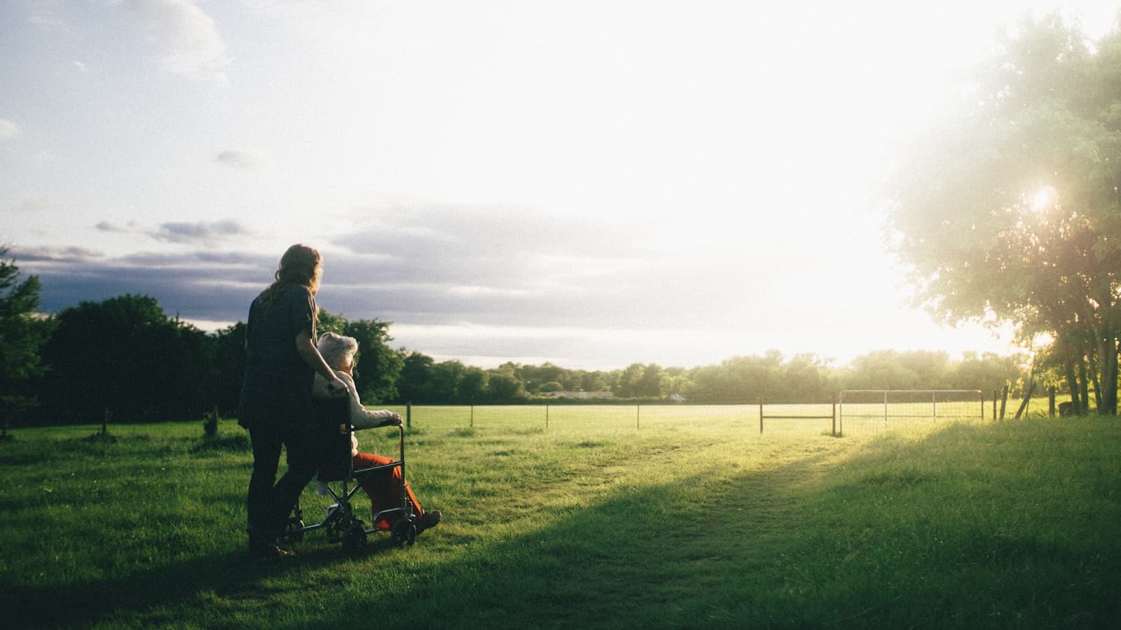 A caregiver walking beside an elderly woman in a wheelchair across a sunlit field.