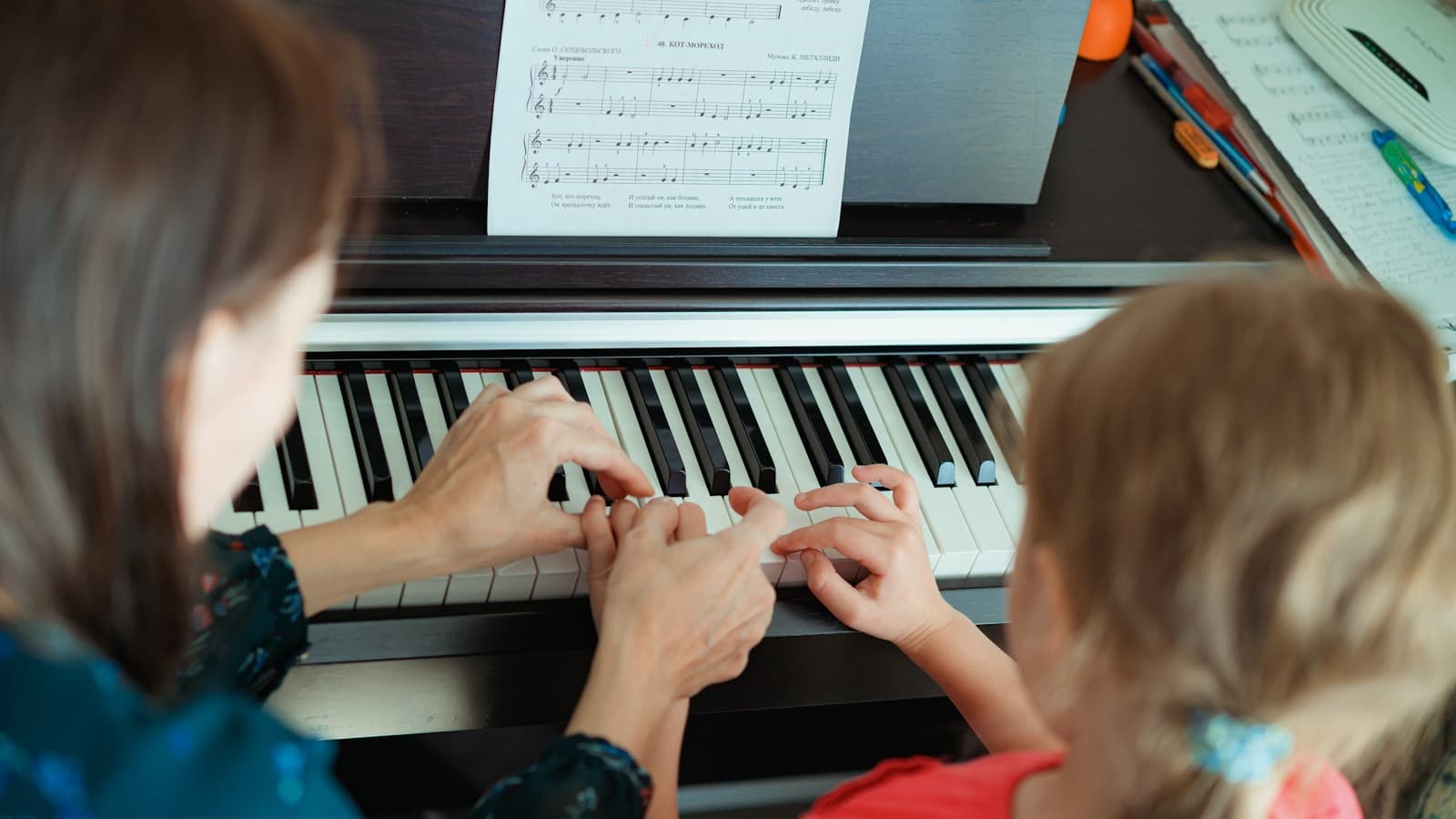 A teacher guiding a child's hands on a piano keyboard, sheet music open.