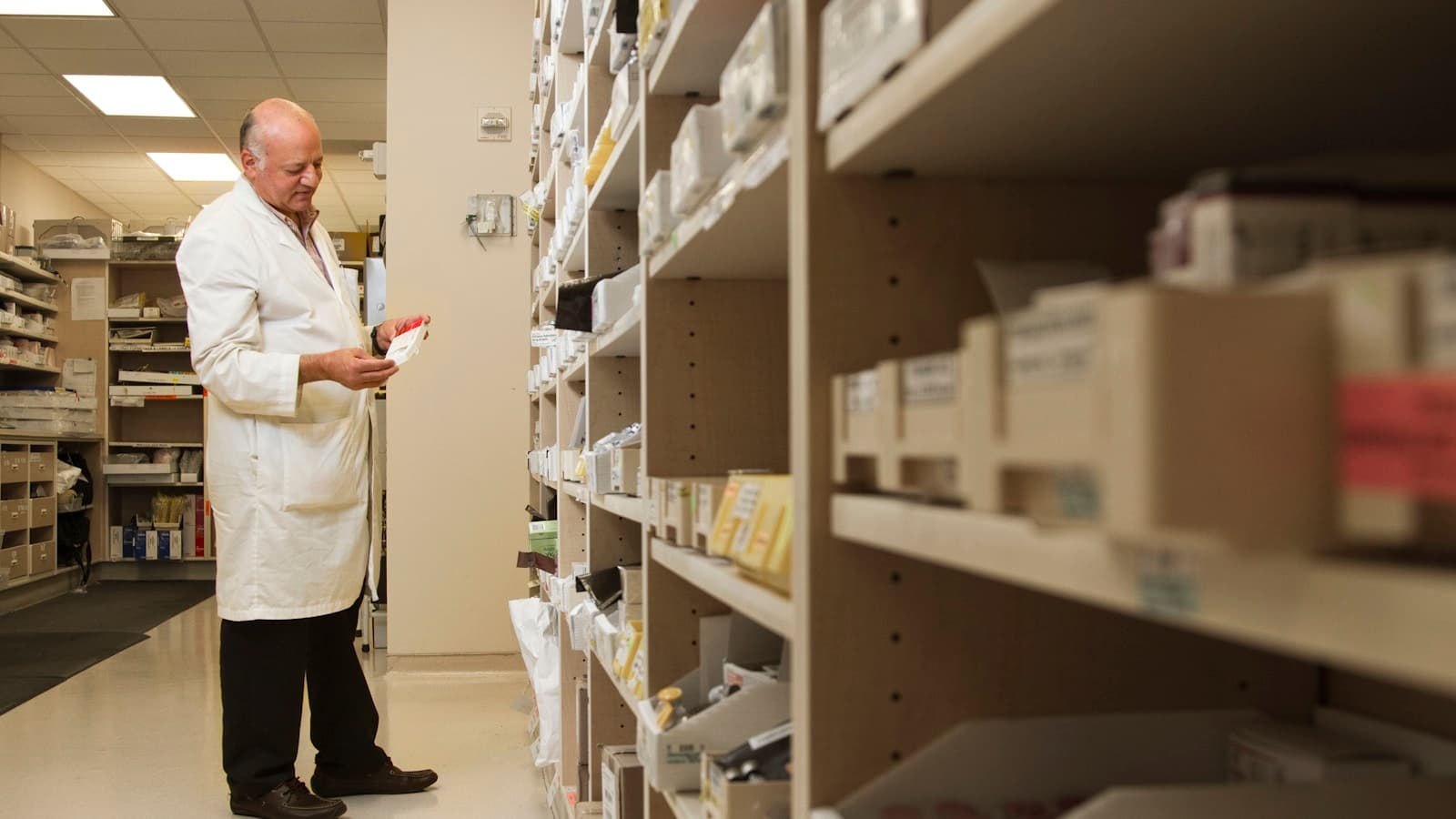 A pharmacist in a white coat examining a small box against a wall of stocked shelves.