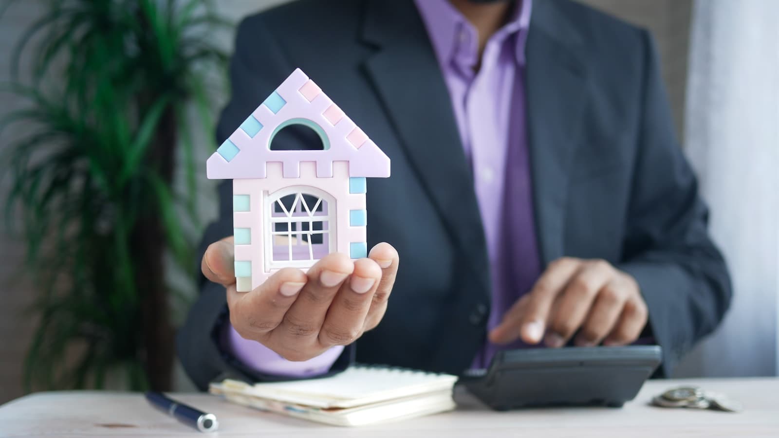 A suited person's torso, face not visible, holding a small toy house model while their other hand rests on a calculator at a desk, no readable text or logos.