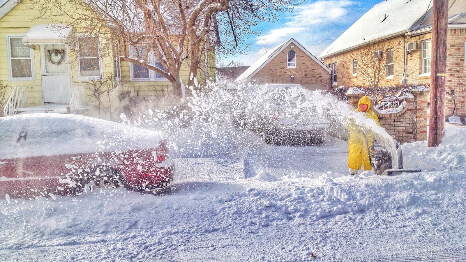 A person in yellow coveralls running a snow blower on a driveway between houses.