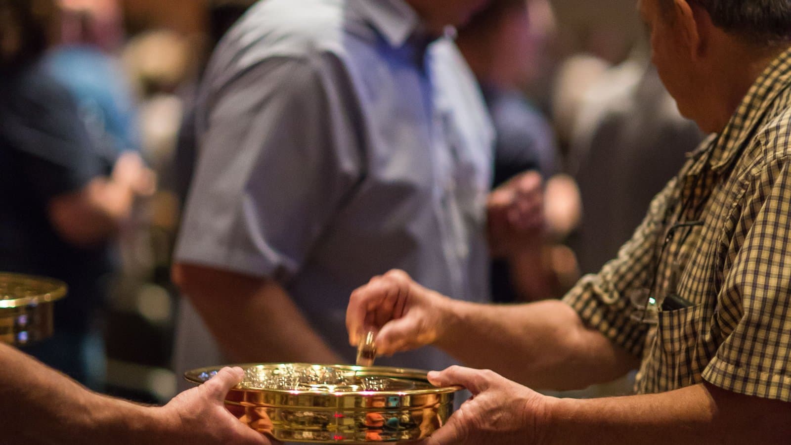 A gold offering plate being passed hand-to-hand between two people seated in a church pew.
