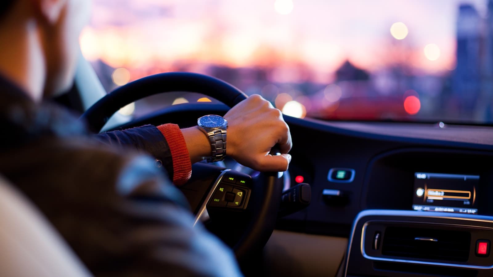 A driver's hand on the steering wheel at dusk with city lights through the windshield.