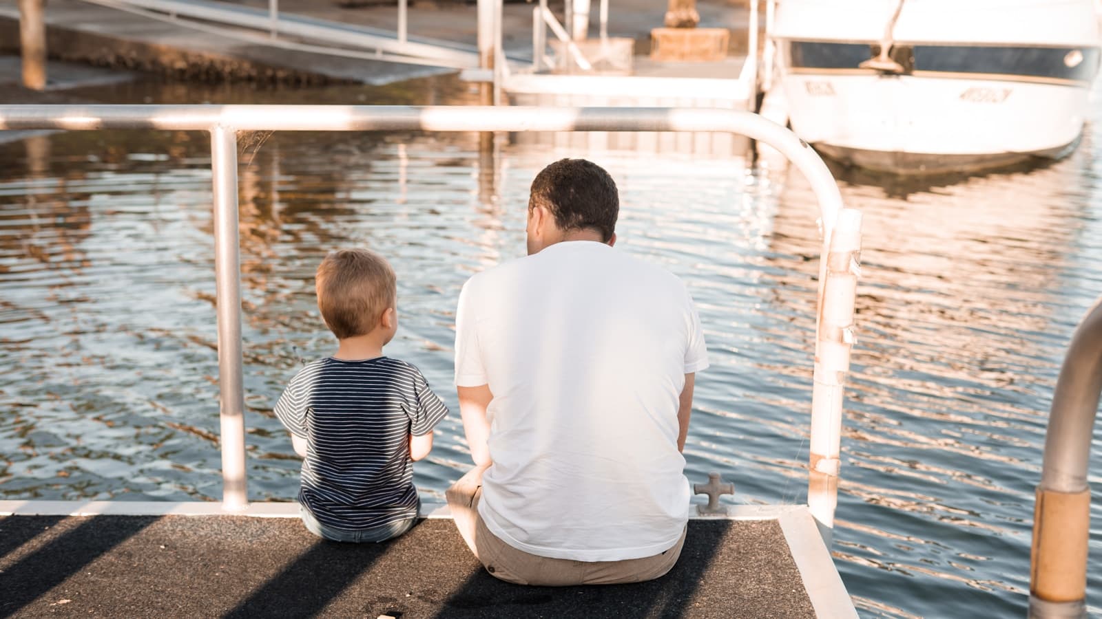A father and son sitting together on a dock looking out at boats.