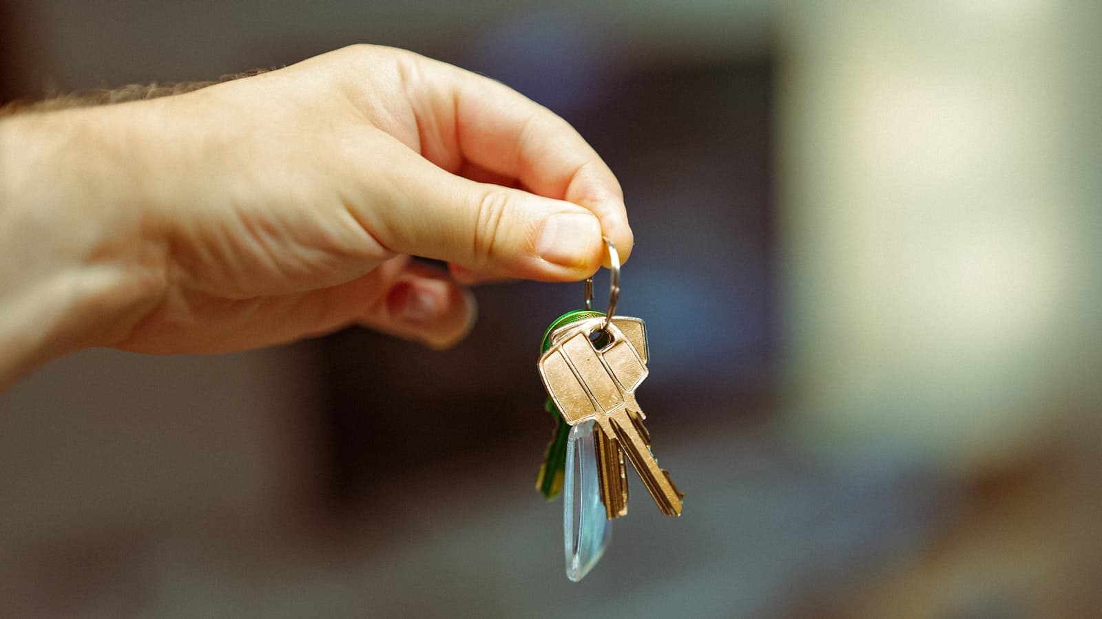 A close-up of a single hand holding up a small set of house keys against a softly blurred indoor background - no face, text, or logos visible.