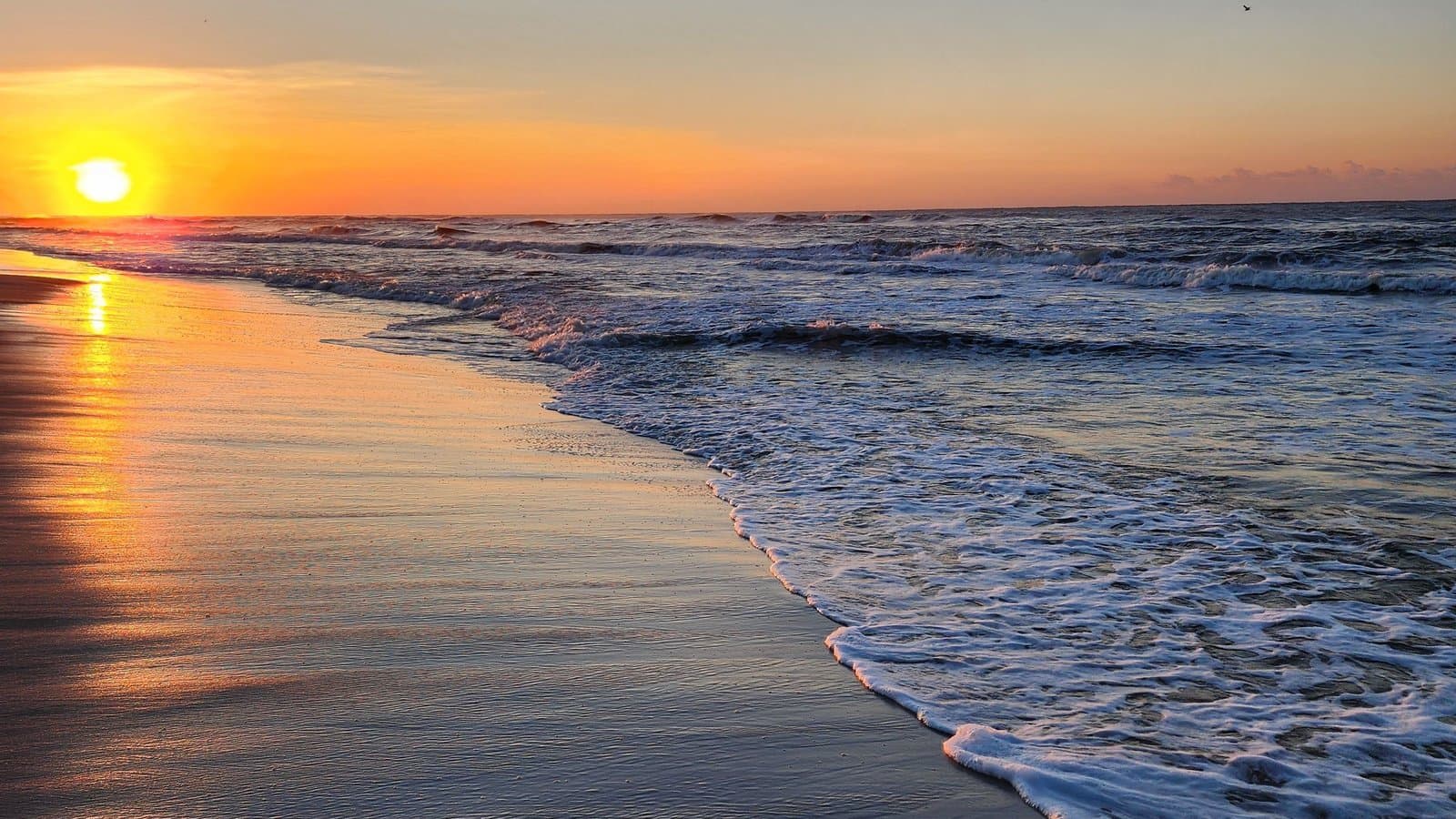 A golden sunset over the Gulf Coast shoreline near Gulf Shores, Alabama, with waves washing onto the beach.