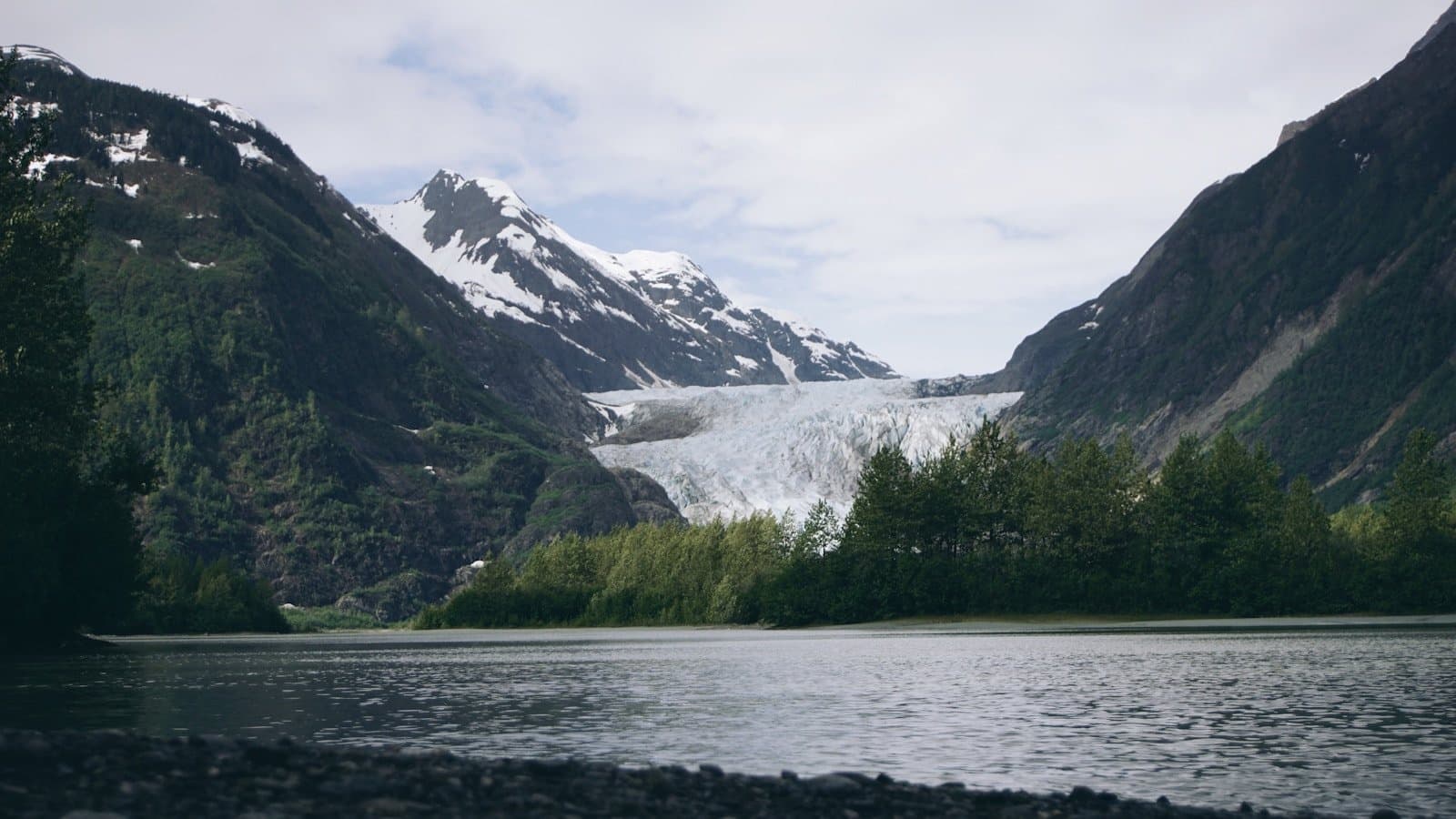 A glacier spilling down between forested mountains into a calm lake.