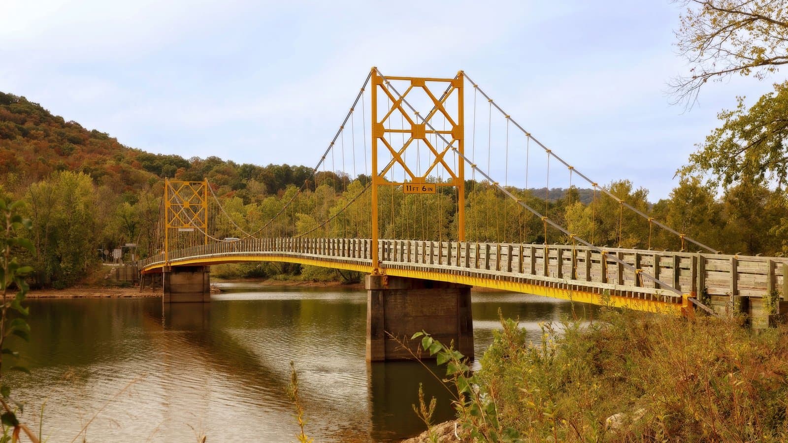 The historic yellow Beaver Bridge, a one-lane suspension bridge over the White River near Eureka Springs, Arkansas, with an autumn hillside behind it.