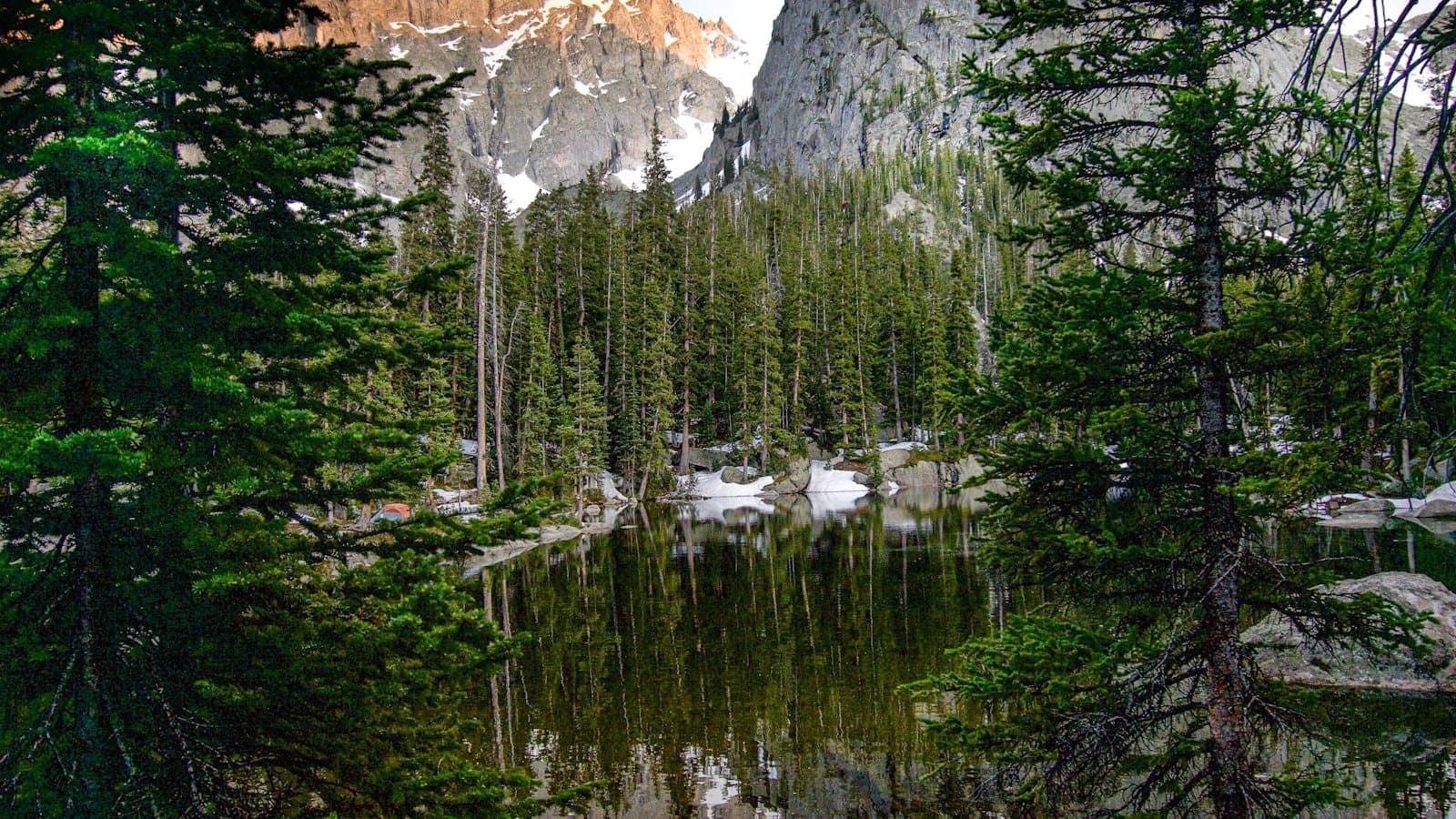 A jagged granite peak reflected in a calm alpine lake ringed by pine trees in the Rocky Mountains.