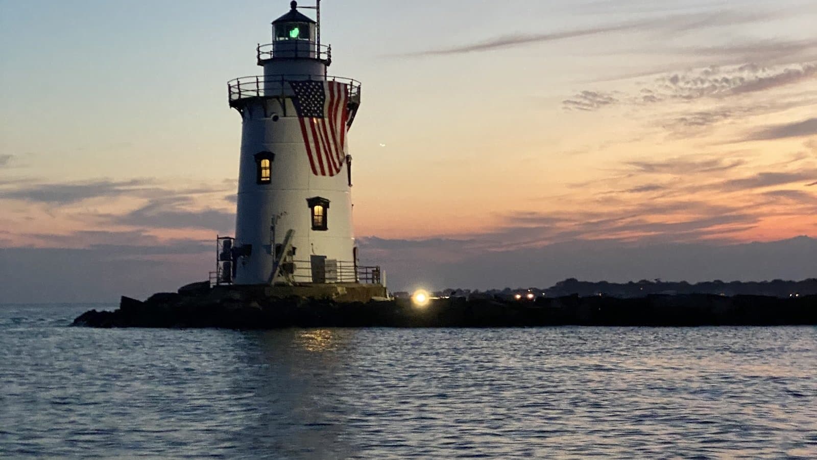 A white New England lighthouse flying an American flag at sunset over calm water.