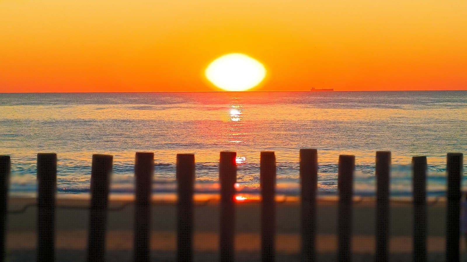 A vivid orange sunrise over the ocean, viewed through the silhouette of a wooden snow fence on a Delaware beach.