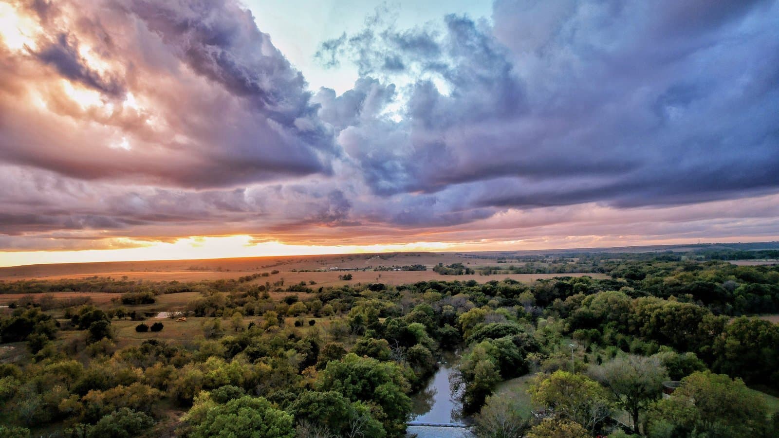 A dramatic aerial sunset over rolling green Flint Hills prairie and a tree-lined creek near Eureka, Kansas.