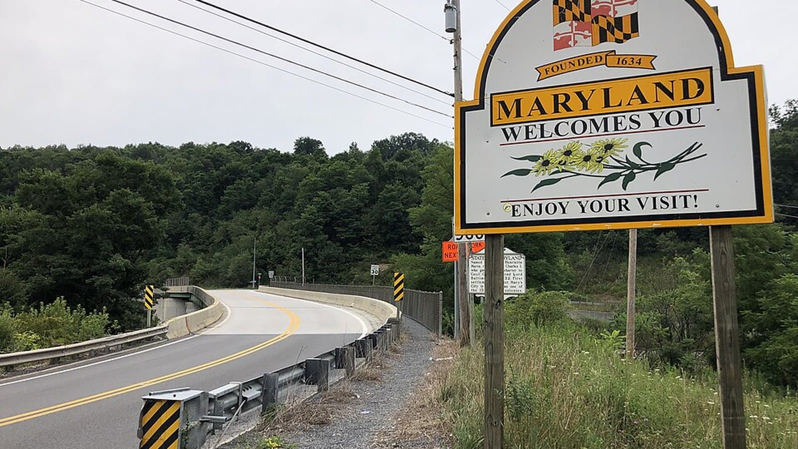 A "Maryland Welcomes You - Founded 1634" highway sign with the state flag crest, next to a rural mountain road.