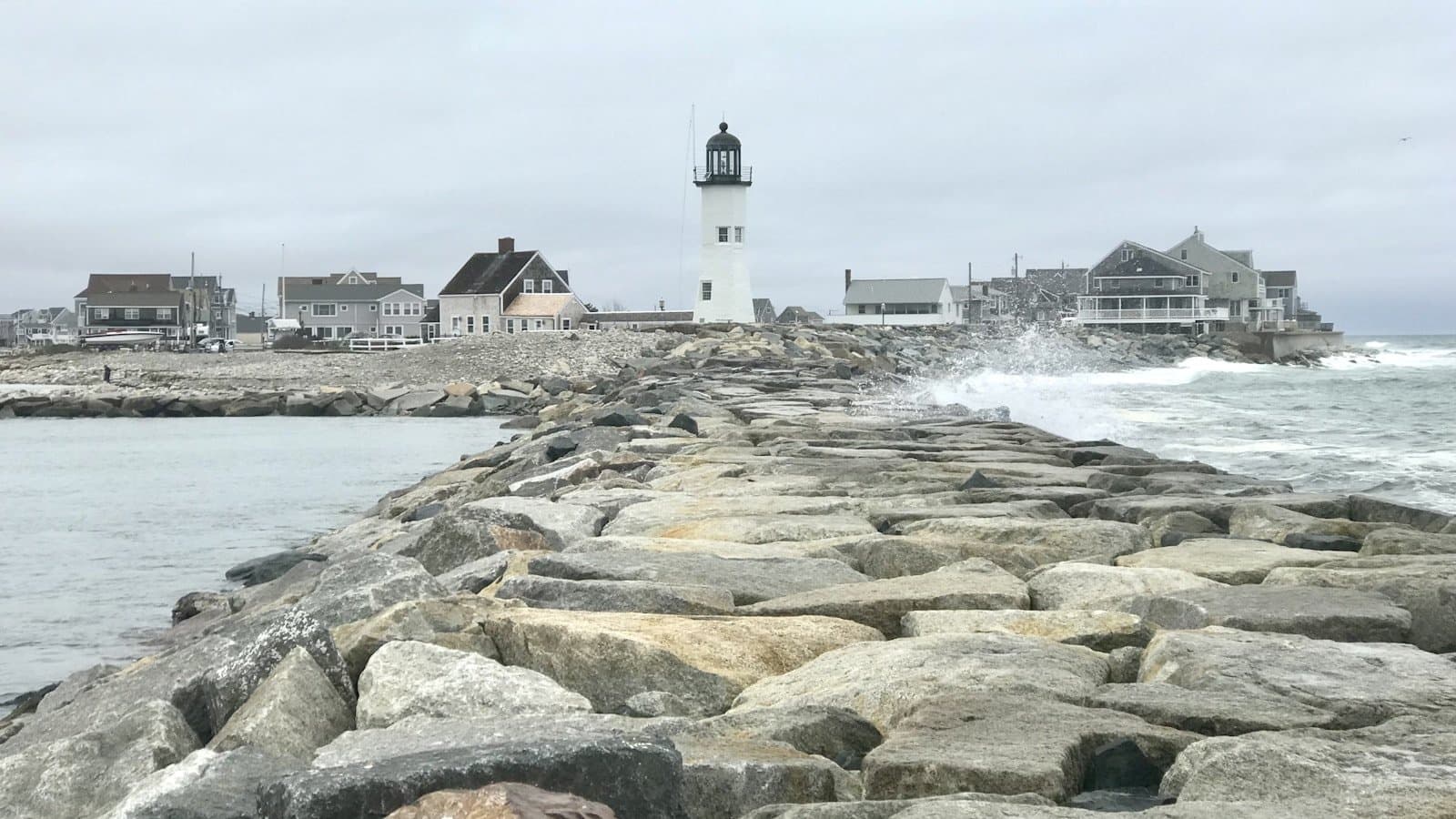 Scituate Lighthouse at the end of a rocky stone jetty on the Massachusetts coast, waves crashing against the rocks.