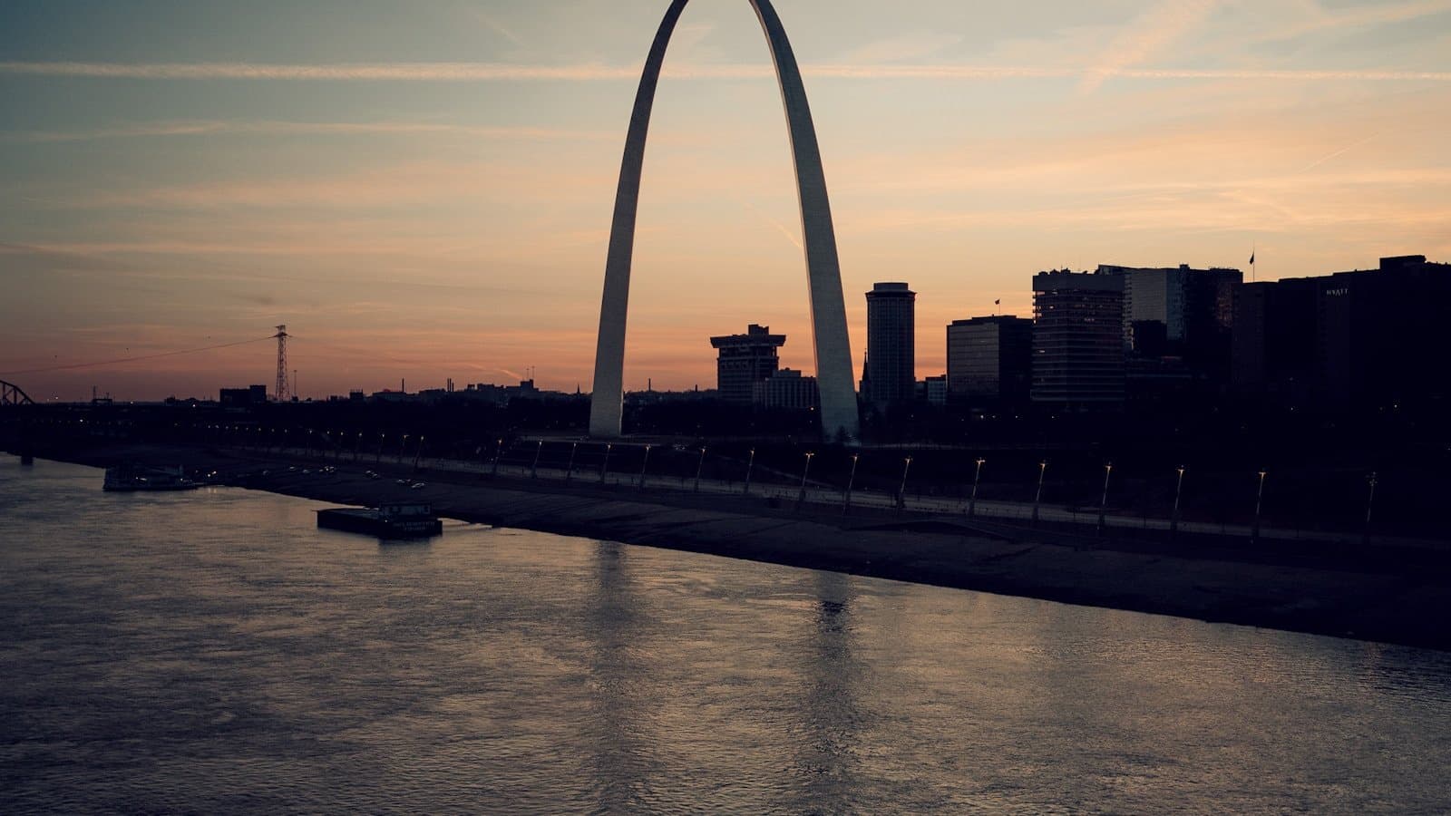 The Gateway Arch silhouetted against a sunset sky above the Mississippi River in St. Louis, with a barge passing below.