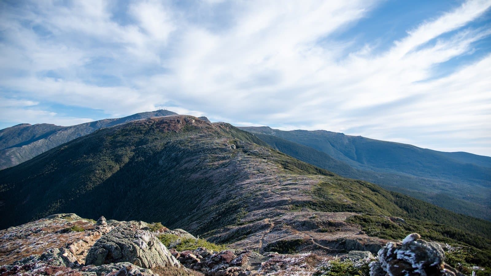 A rocky mountain ridge trail overlooking layered peaks of the White Mountains, New Hampshire, under a partly cloudy sky.