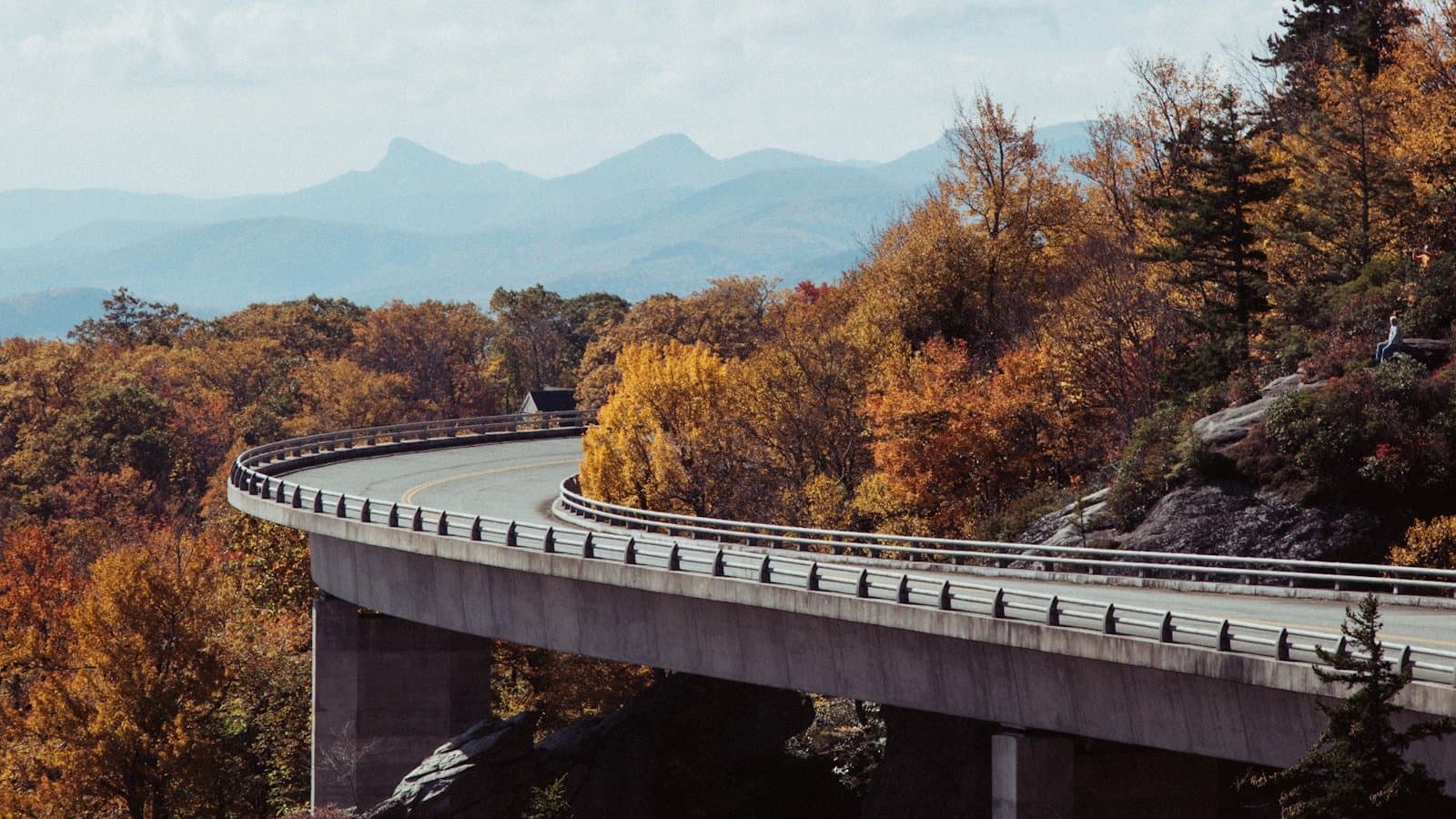 The Linn Cove Viaduct curving along the Blue Ridge Parkway through peak autumn foliage with mountains in the background.