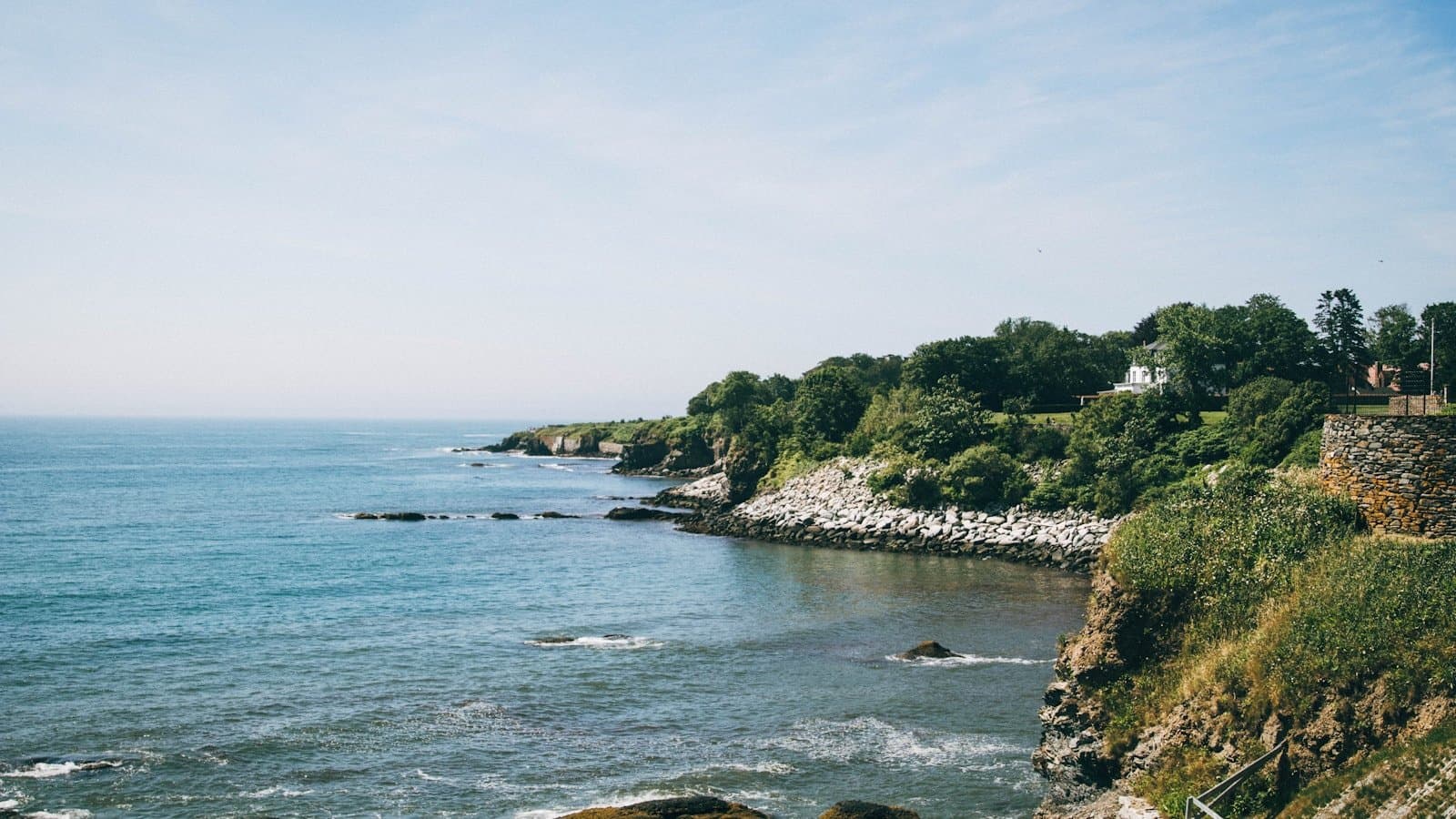 The rocky Newport, Rhode Island coastline and Cliff Walk area with blue ocean, green shrubs, and a stone wall.